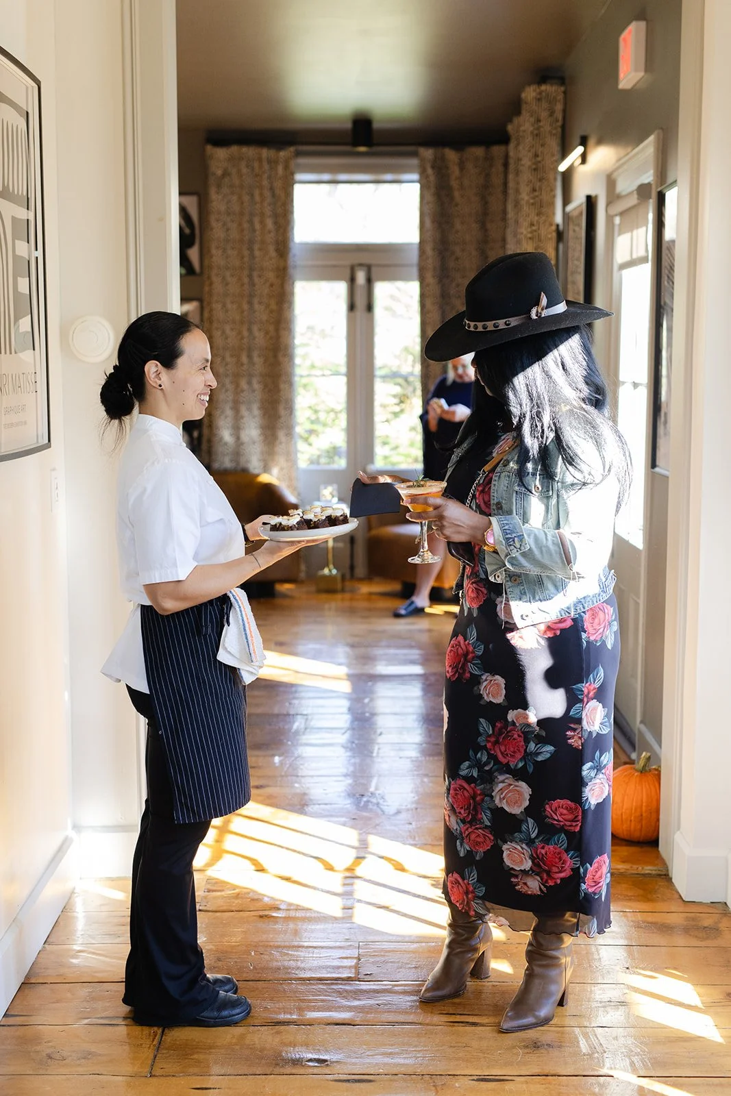 A woman in a white shirt and striped apron serving desserts to a woman in a floral dress and hat at a social gathering indoors with sunlight streaming in.