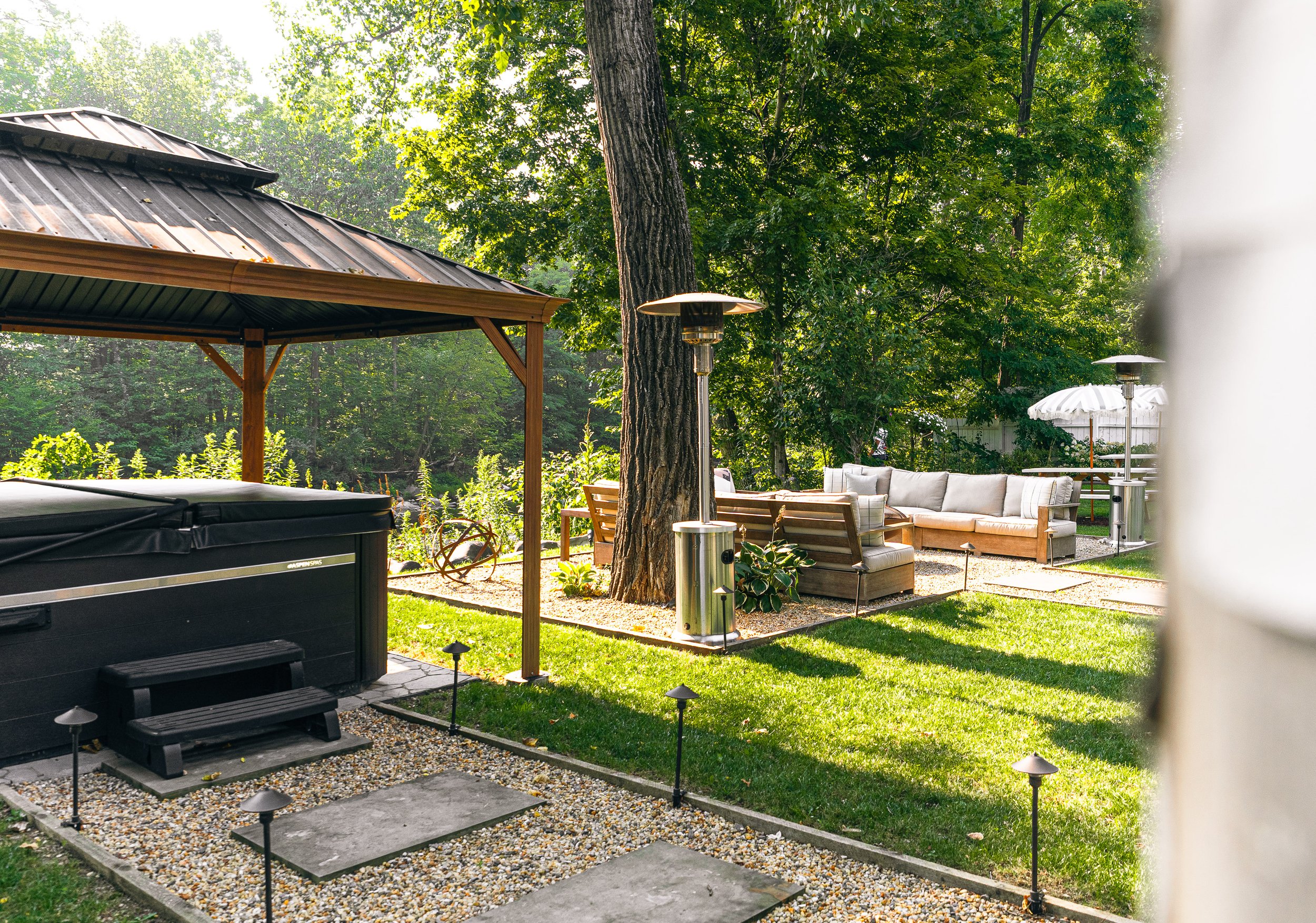Outdoor backyard with seating area, large tree, hot tub, patio heaters, gazebo, and garden in the background.