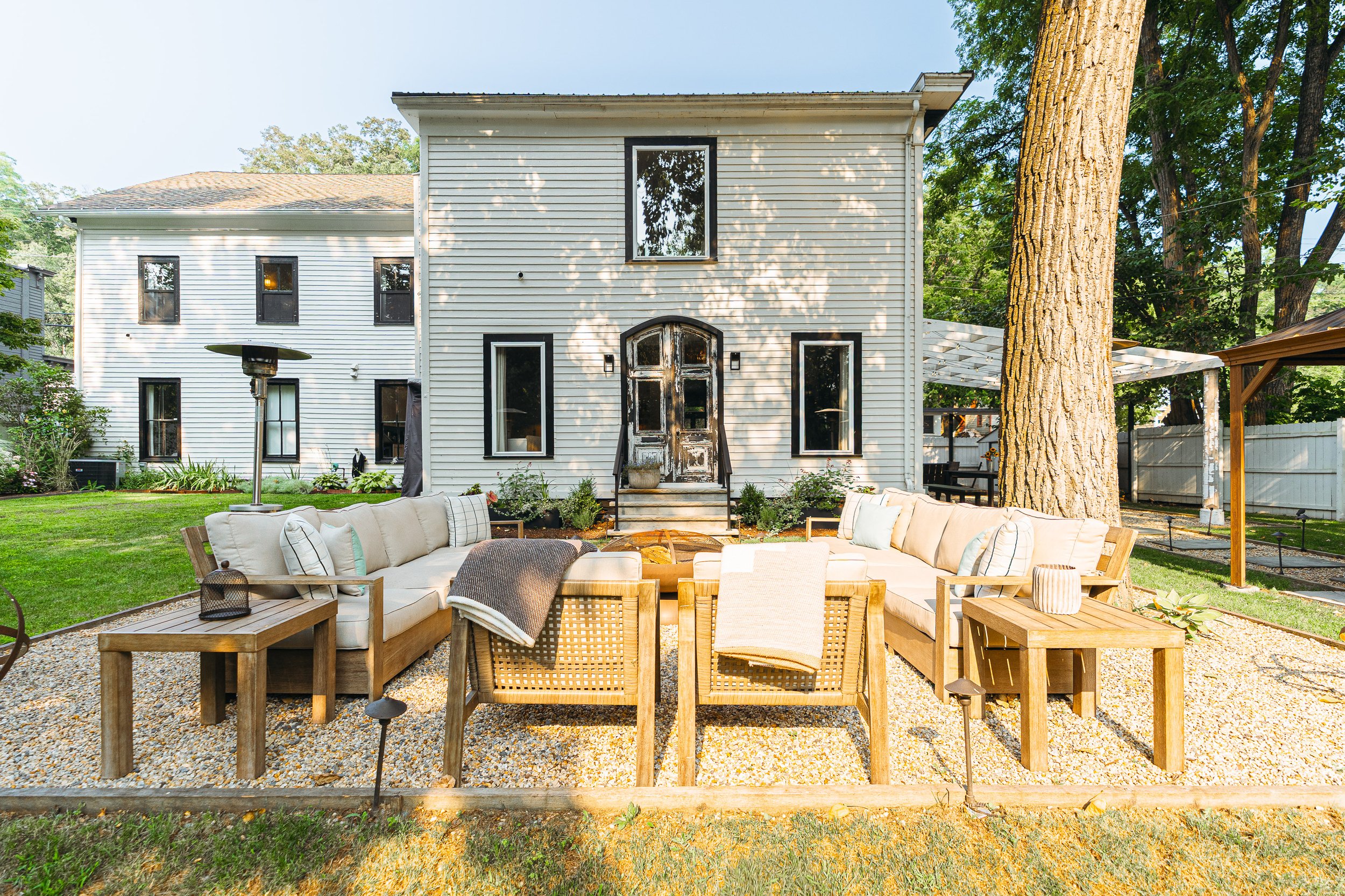 Backyard patio with outdoor seating including beige cushioned sectional sofas and wooden tables in front of a white house with black window frames, large trees, and a pergola.