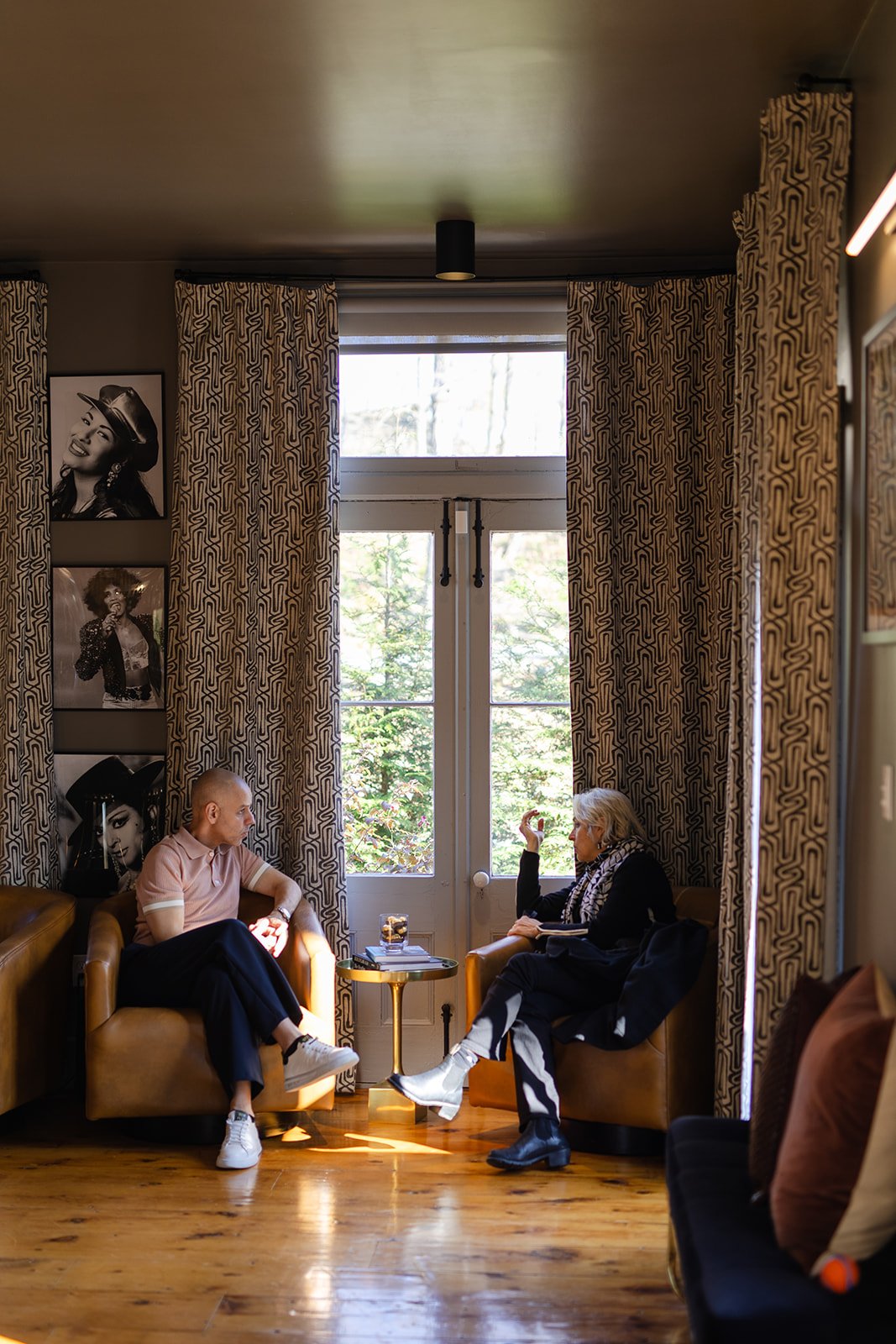 A man and a woman sitting on separate armchairs in a cozy room, engaged in conversation near a window with patterned curtains. There are black-and-white photographs on the wall behind the man, and a small side table with a glass of water and magazines between them.