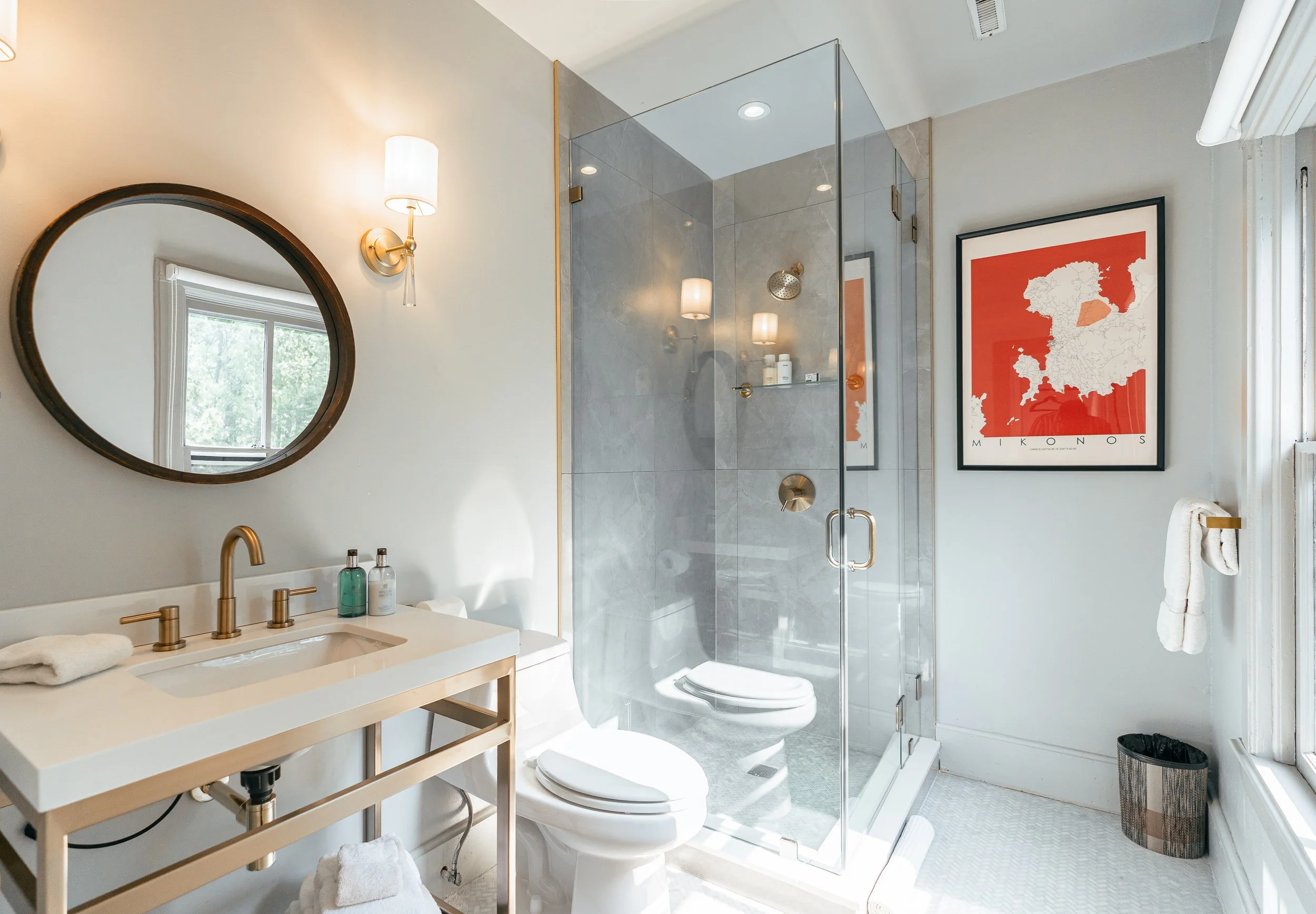 A modern bathroom with a glass shower enclosure, a white toilet, a white vanity with a gold faucet, a round mirror, a wall sconce, framed artwork with a map of Europe, a towel on a hook, and a small wastebasket.