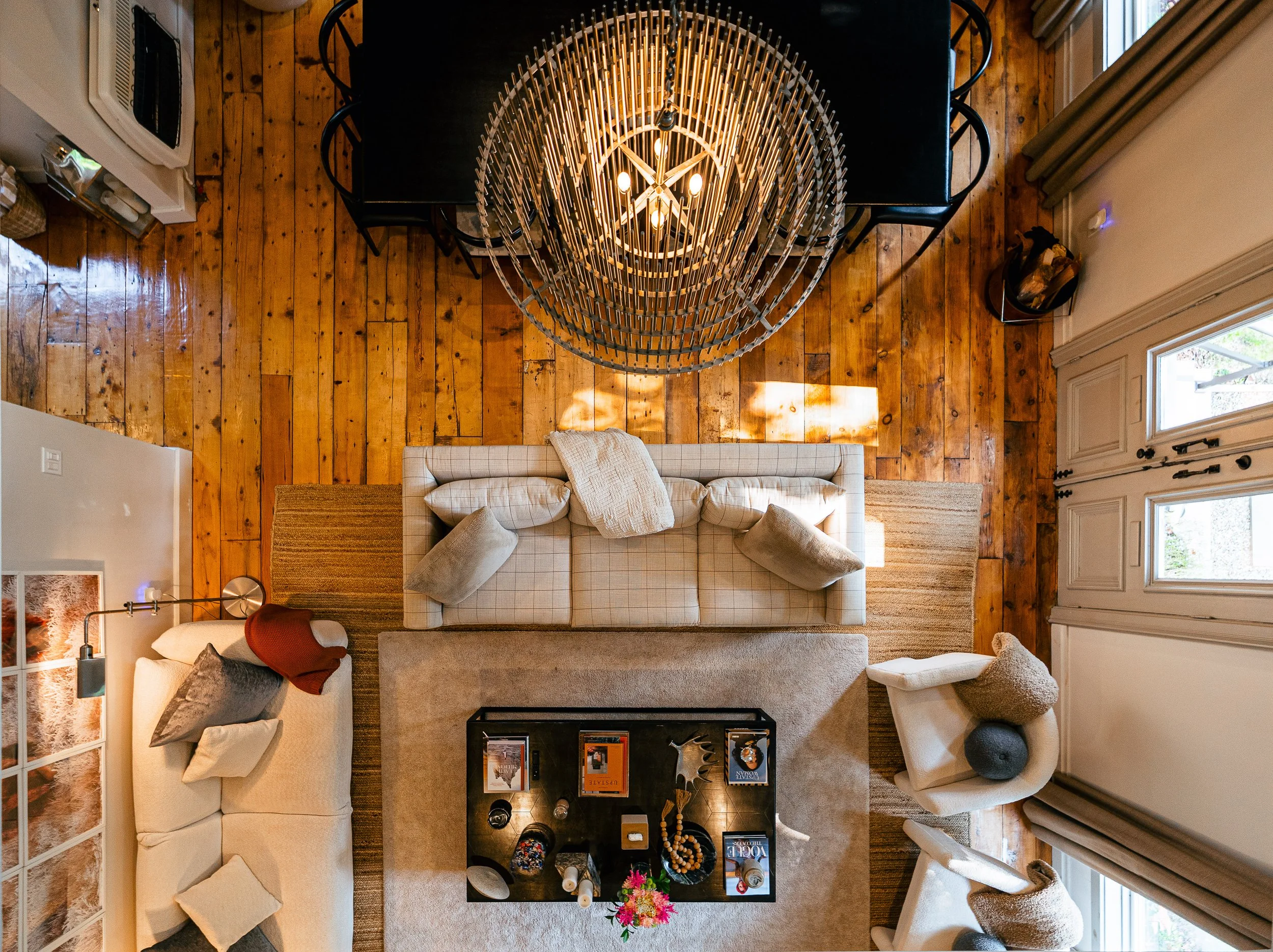 Overhead view of living room with wooden floor, beige sofas, armchairs, coffee table, and a black dining table with chairs, illuminated by a chandelier.
