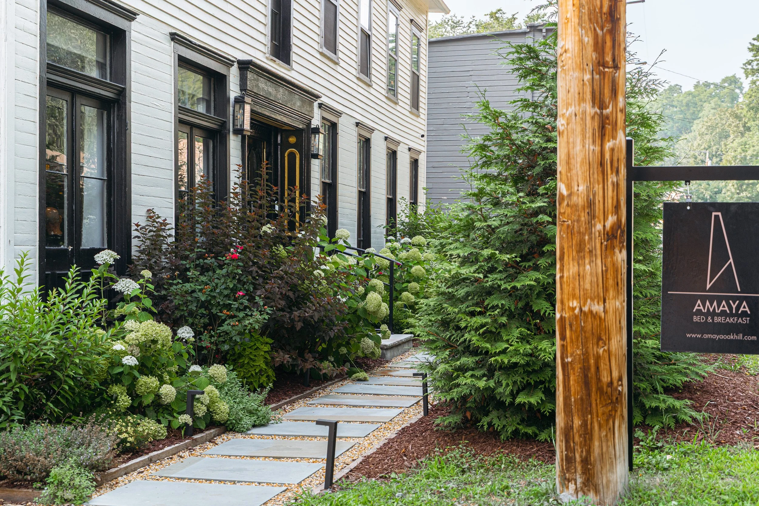 A pathway with stepping stones leading to a white house with black window frames and a black door, surrounded by greenery and flowering plants, with a wooden utility pole on the right and a sign for Amaya Bed & Breakfast.