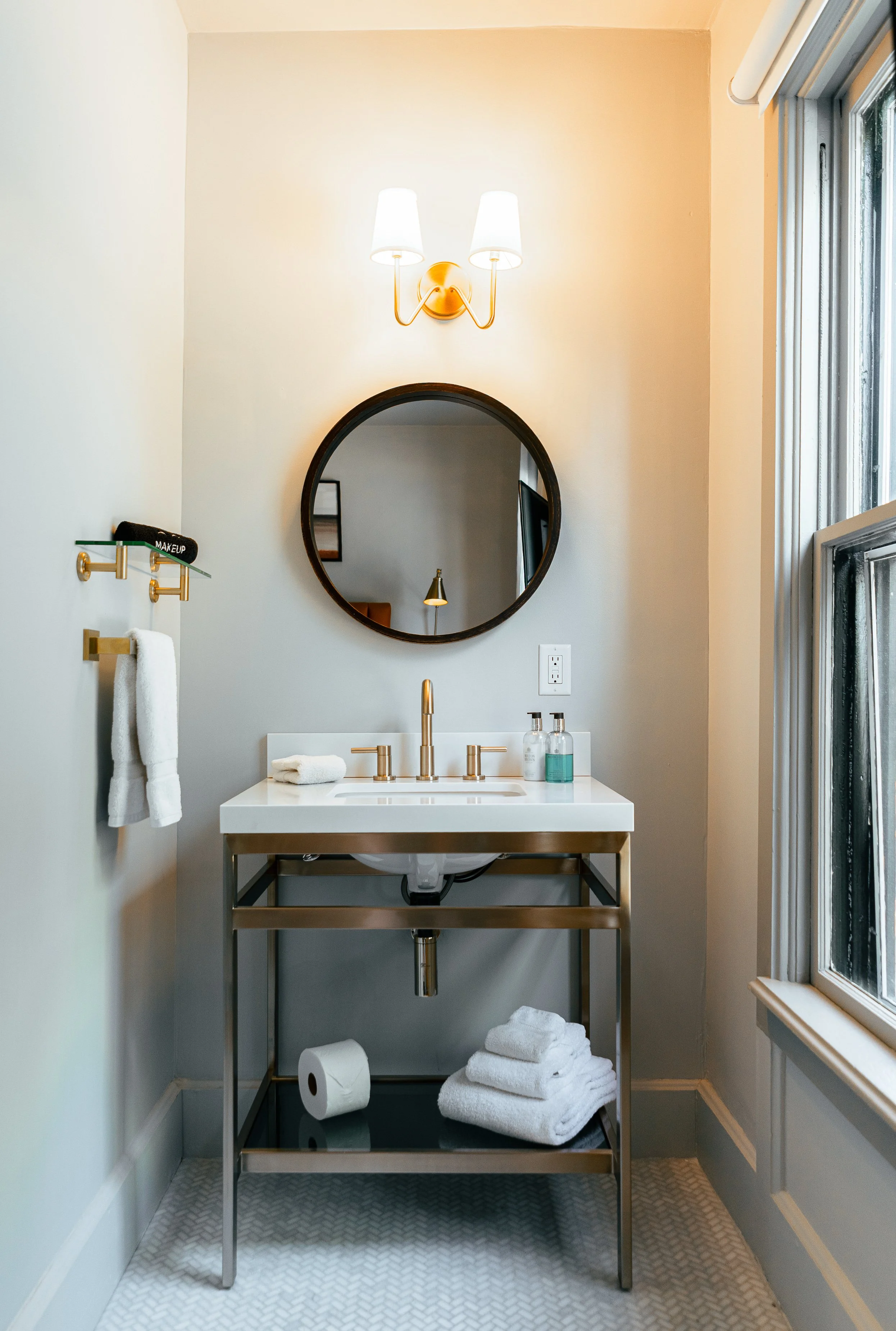 Small bathroom with a white sink on a metal stand, a round mirror, a wall sconce with twin lamp shades, a towel rack with a towel and makeup brush, and a window with natural light.