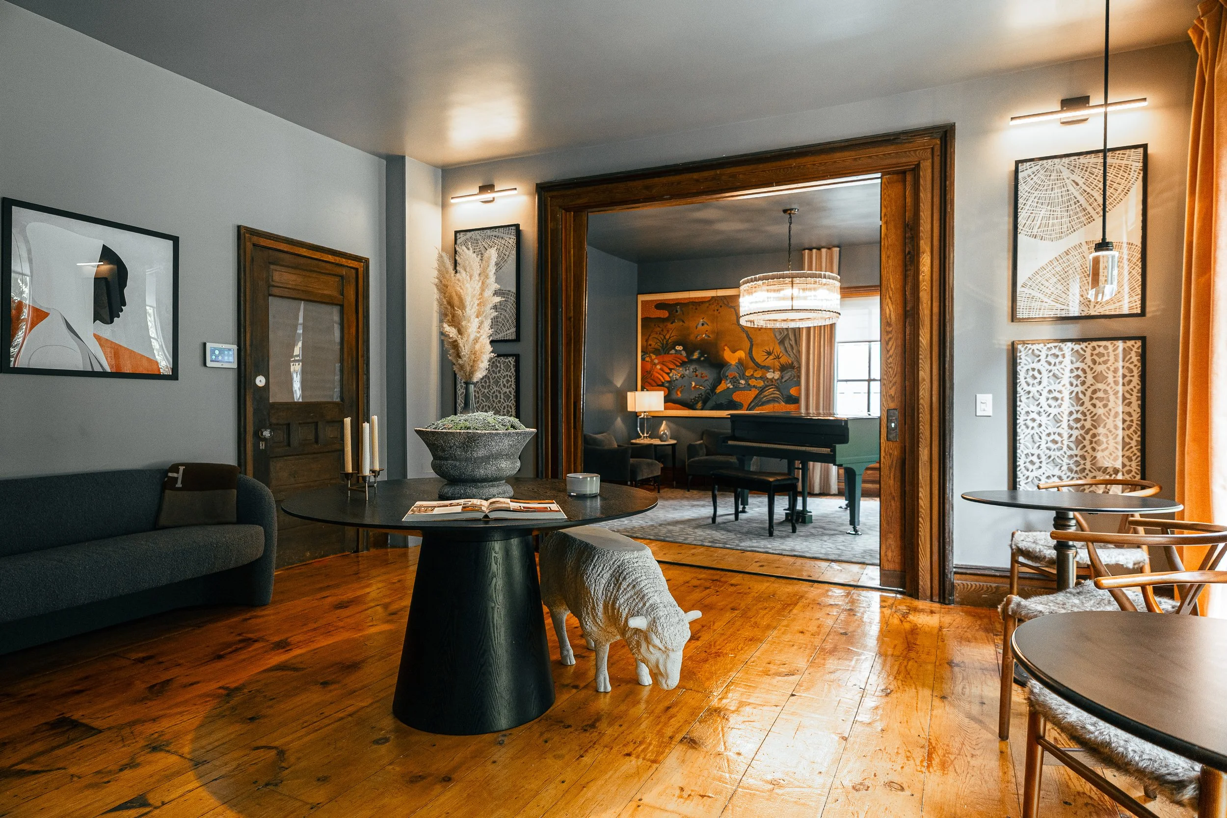 Living room with wooden floors, gray walls, and modern artwork. Contains a black couch, round black dining table with a sheep sculpture underneath, and a large stone vase with pampas grass. An open doorway leads to a sitting area with a grand piano, wall art, and a chandelier.
