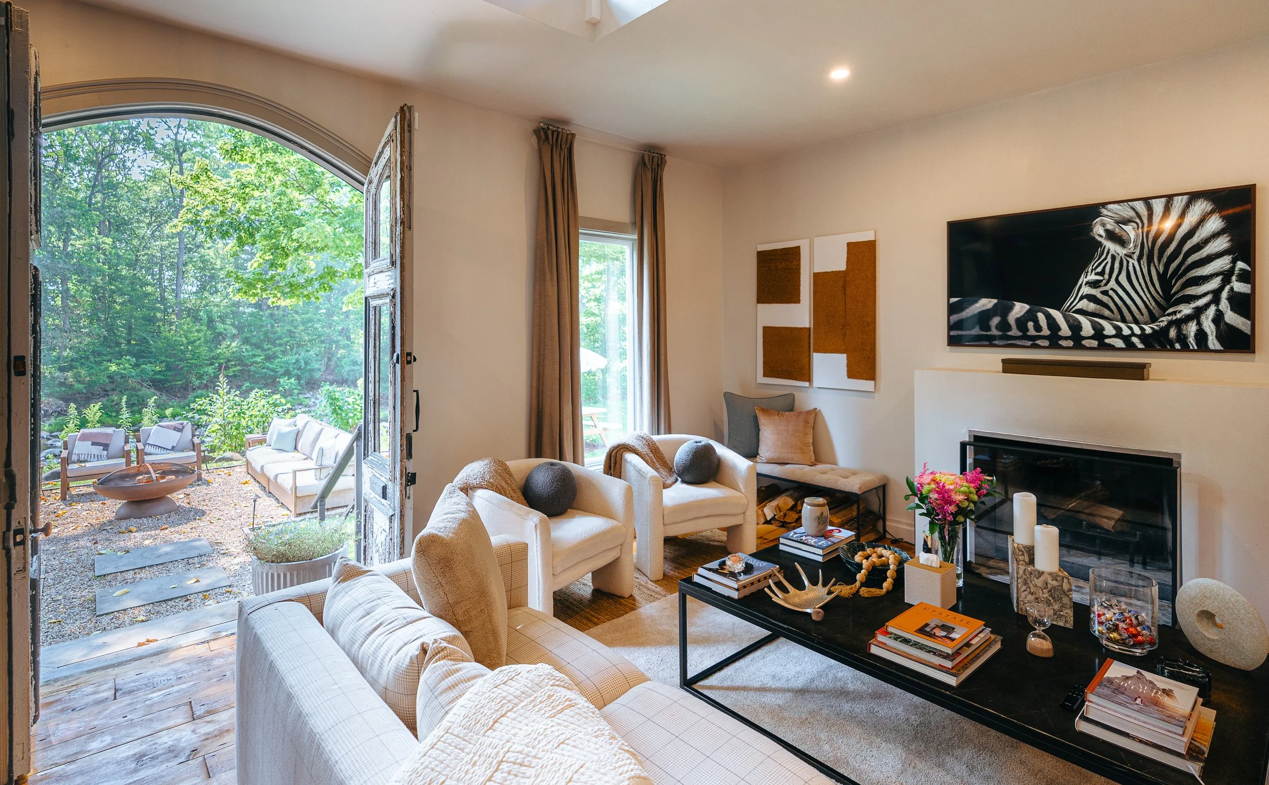 Living room with beige sofa, armchairs, coffee table with books, candles, and decorative items, indoor fireplace, wall art, and open door leading to outdoor patio with chairs, trees, and firepit.