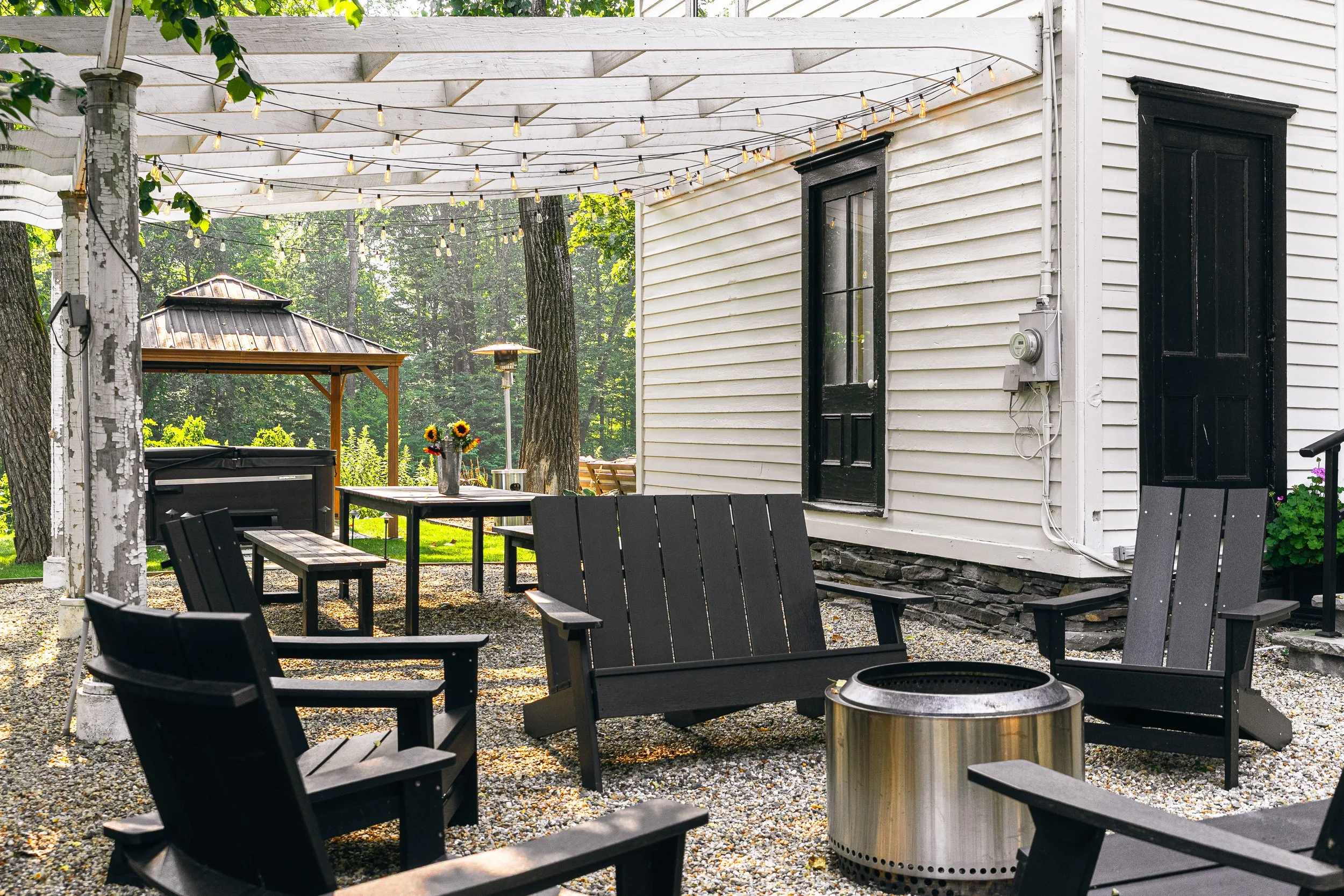 Backyard patio with black Adirondack chairs, fire pit, floral arrangements, string lights overhead, small gazebo in the background, and a white house with black doors.
