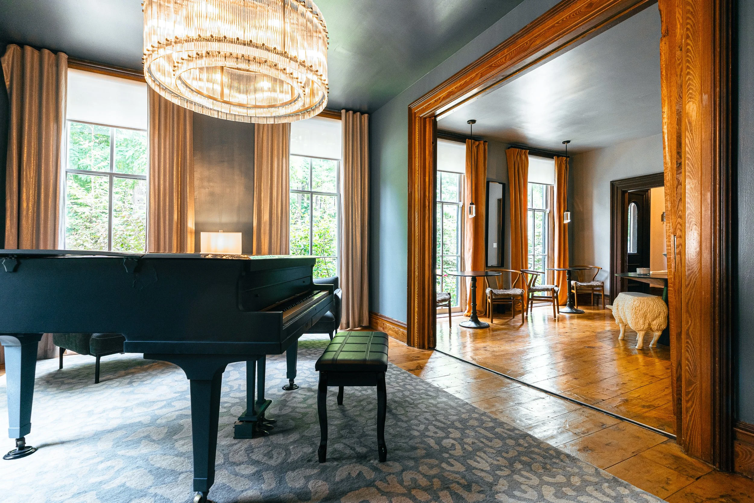 A spacious room with a black grand piano on a patterned rug, large windows with beige curtains, a chandelier overhead, and a separate sitting area with wooden floor, tables, and chairs.