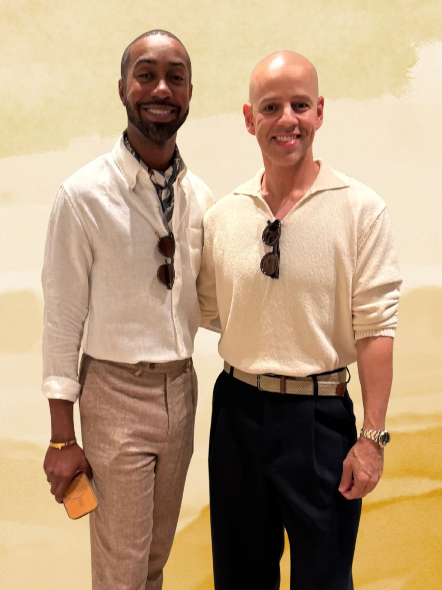 Two smiling men standing together indoors, one with dark skin and the other with light skin, both wearing light-colored shirts and sunglasses hanging from their shirts.