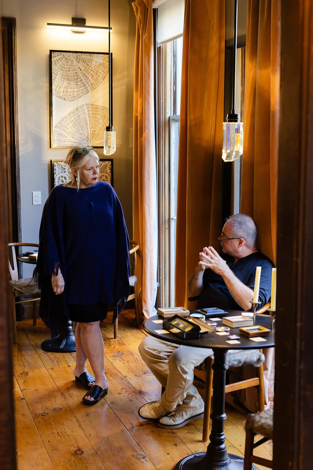 A woman with blonde hair, wearing a blue poncho and sandals, is standing and talking to a man with glasses, wearing a black shirt and beige pants, who is seated at a round table in a cozy room with wooden floors and orange curtains.