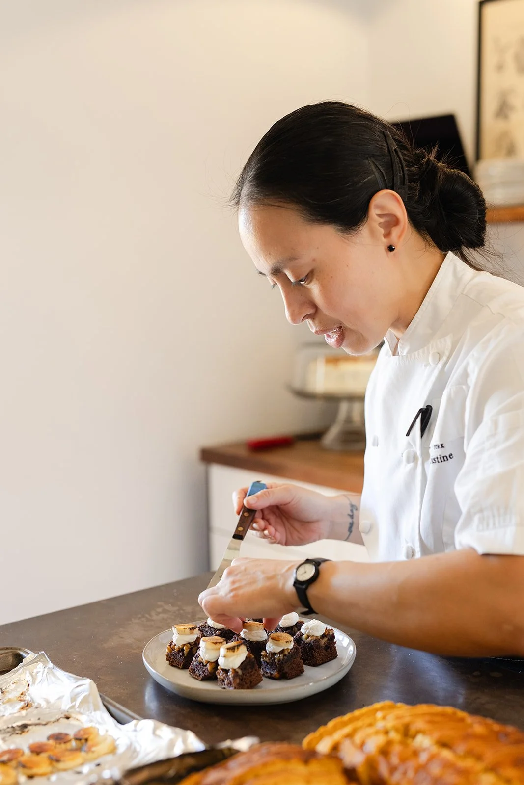 A woman in a white chef's coat is decorating small chocolate desserts with white cream on a countertop.
