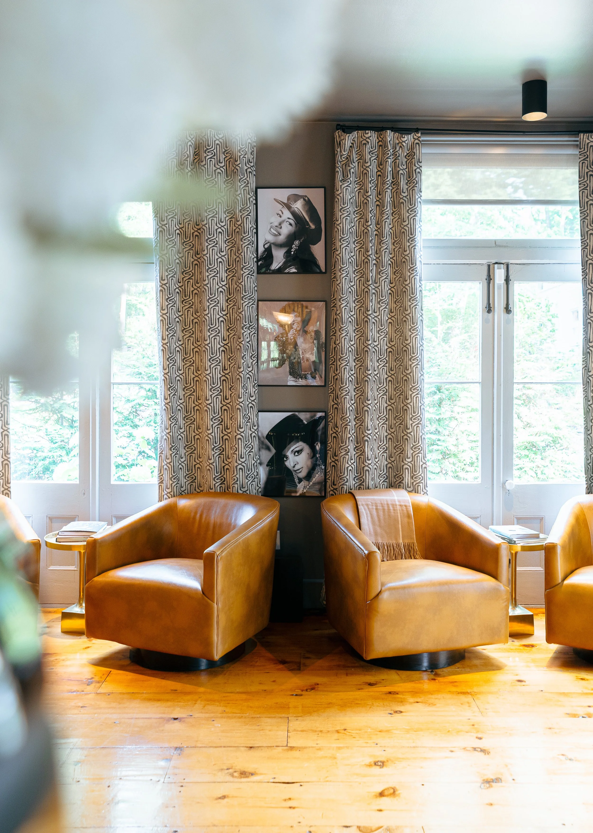 A cozy living room with two mustard-yellow leather armchairs, wooden floors, windows with patterned curtains, and three black-and-white photographs of women on the wall between the windows.