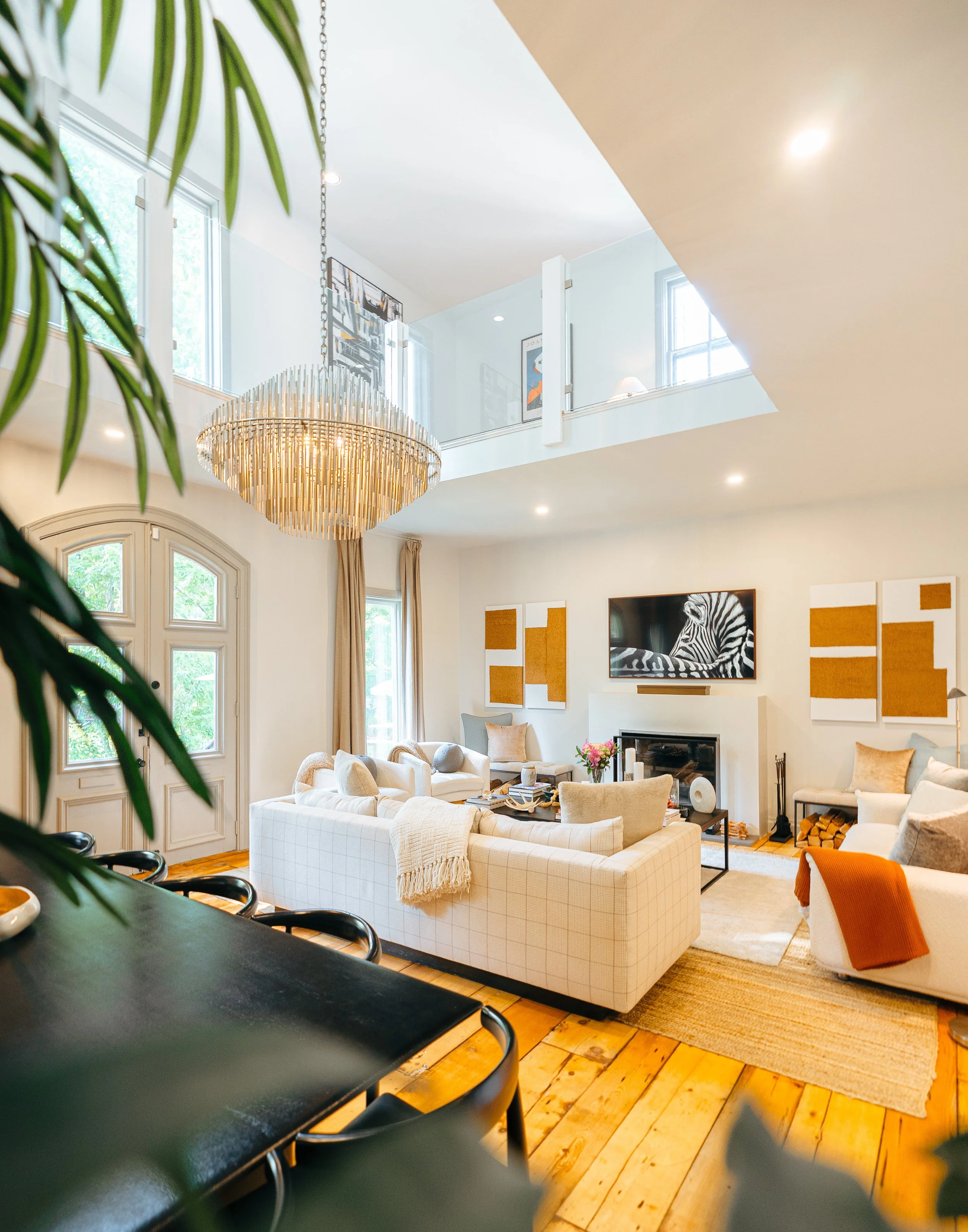 Bright and modern living room with cream-colored furniture, a large chandelier, artwork on the walls, a flat-screen TV with a zebra image, and large windows with beige curtains.