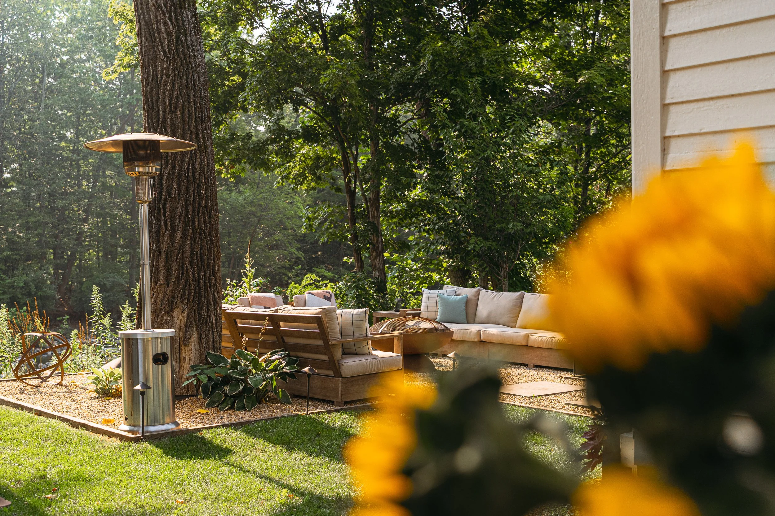 Outdoor patio with beige sofa and chairs, surrounded by green trees and plants, yellow flowers in the foreground, and an outdoor heater to the left.