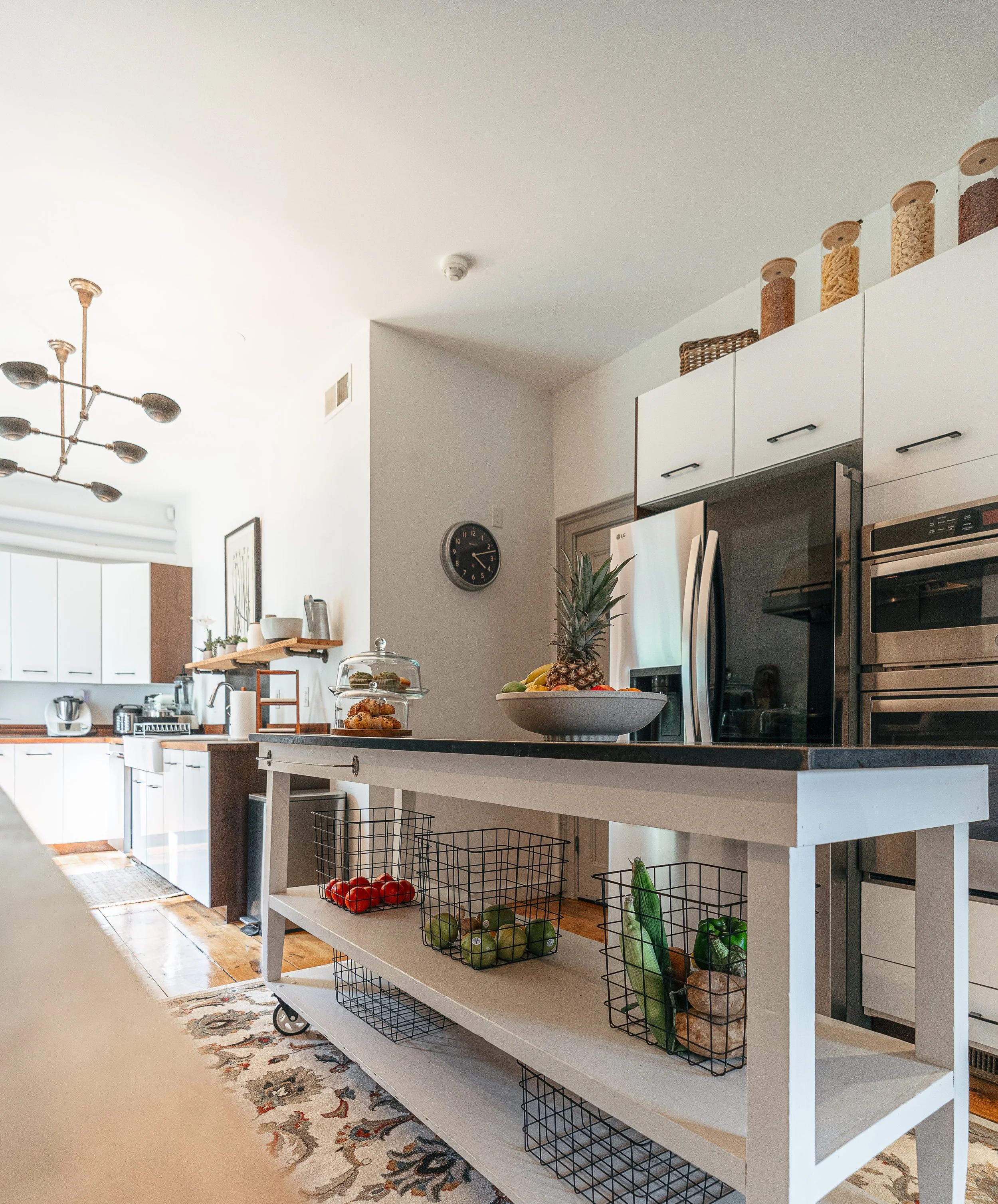Modern kitchen with white and wood cabinetry, black countertops, stainless steel appliances, and open shelving with jars of food on top. A white kitchen island holds a bowl of fruit and a cake stand with baked goods, with wire baskets of vegetables and fruits underneath. A patterned rug covers the wooden floor.