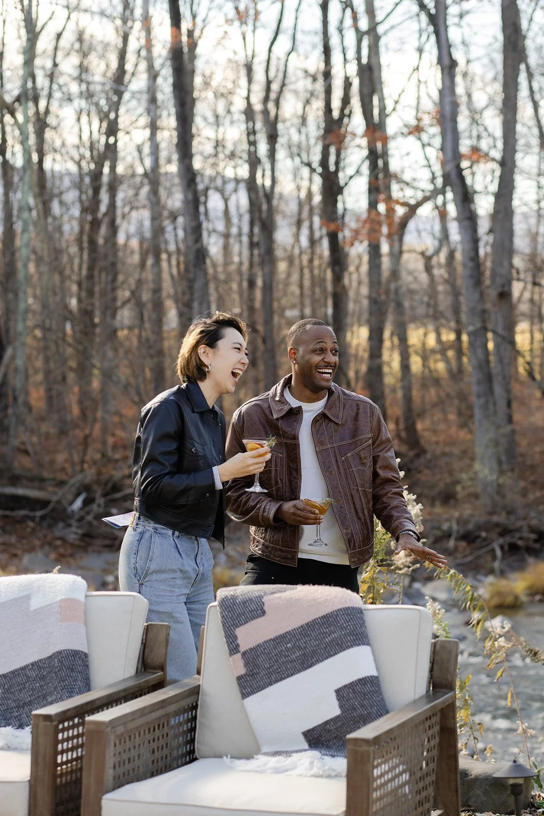 Two people standing outdoors near a wooded area, laughing and holding drinks, with chairs and blankets in the foreground.