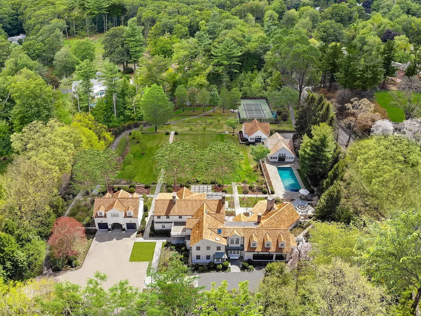 Aerial view of a large estate with multiple houses, a pool, tennis court, and extensive green lawn surrounded by trees.