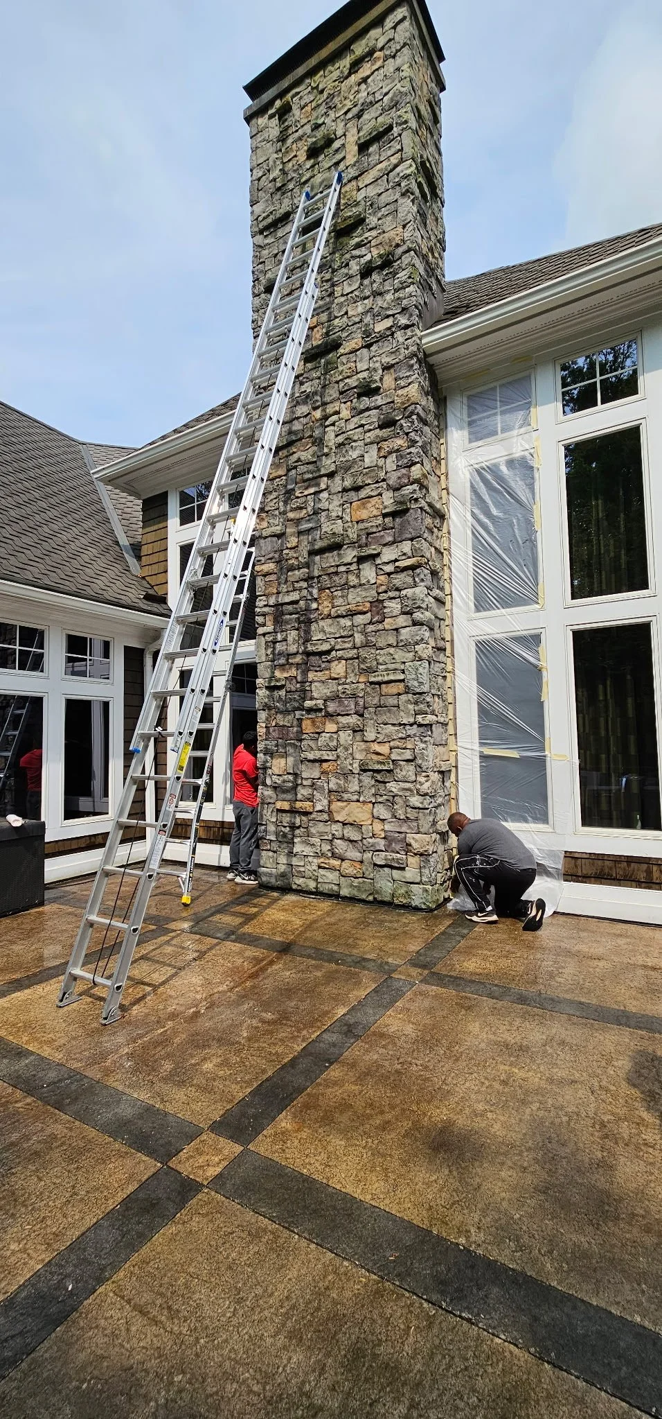 Two men working on the base of a stone chimney in a backyard, with a tall ladder leaning against the chimney, and a large house with multiple windows and a deck in the background.