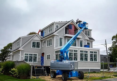 Construction workers on a boom lift working on the upper floors of a large gray house with many windows. The house is surrounded by a yard with green bushes, and there is a partly cloudy sky above.