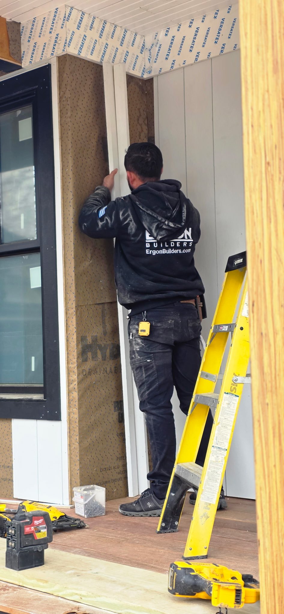 Construction worker installing insulation or paneling on a wall, standing on a wooden floor with tools nearby, ladder beside him in an indoor construction site.