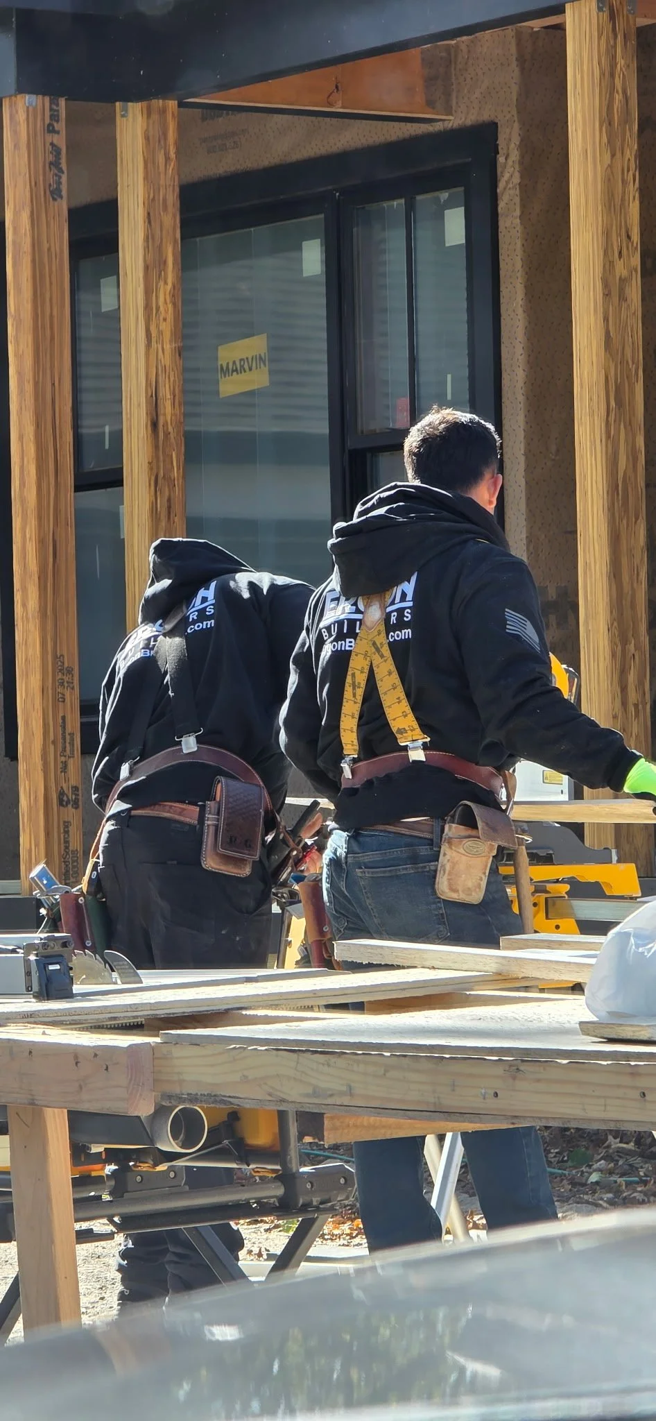 Two construction workers at a building site, wearing black hoodies with logos, with wooden framing and construction tools around them.