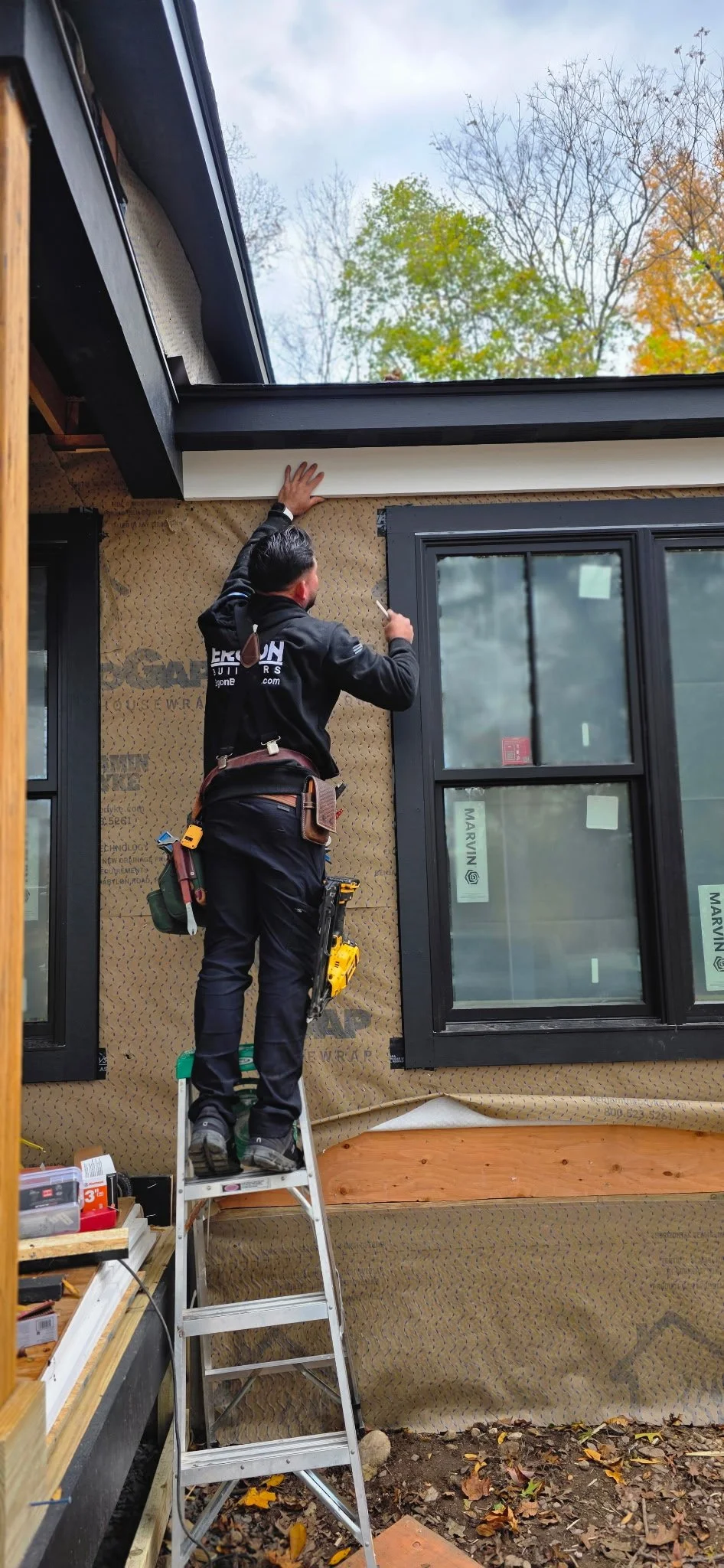 Construction worker standing on a ladder installing or repairing exterior trim around a large window on a house, with fall foliage visible in the background.