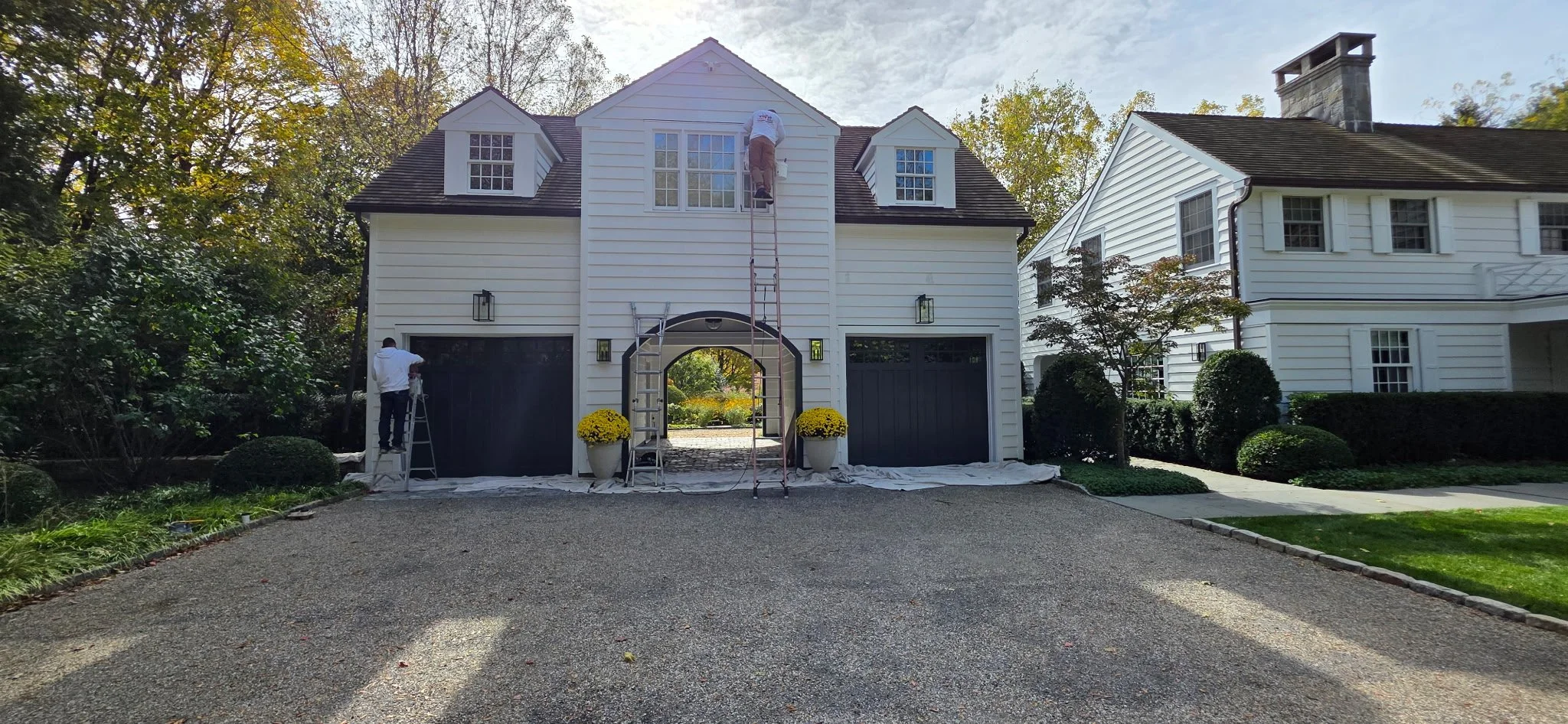 Two workers painting the exterior of a white house with black doors and a small archway entrance, surrounded by greenery and autumn trees.