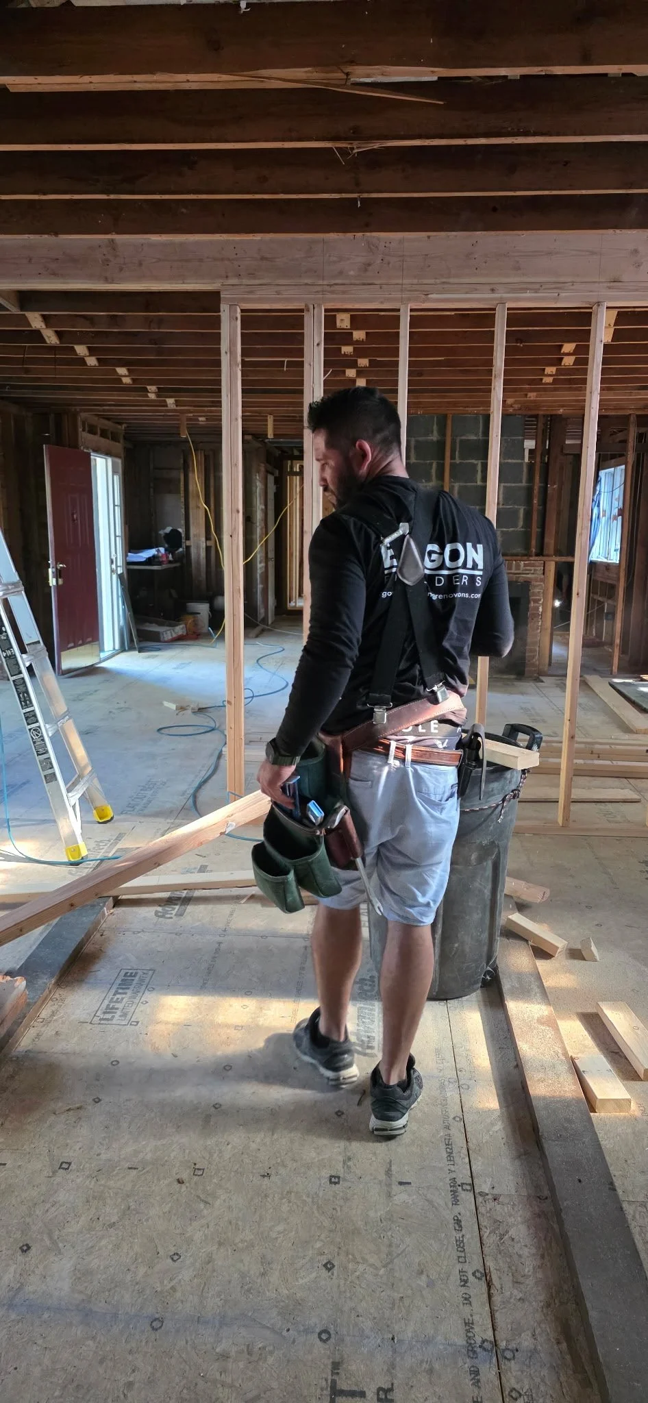 A construction worker in a black shirt and gray shorts standing inside a house under renovation, surrounded by wooden framing and construction tools.