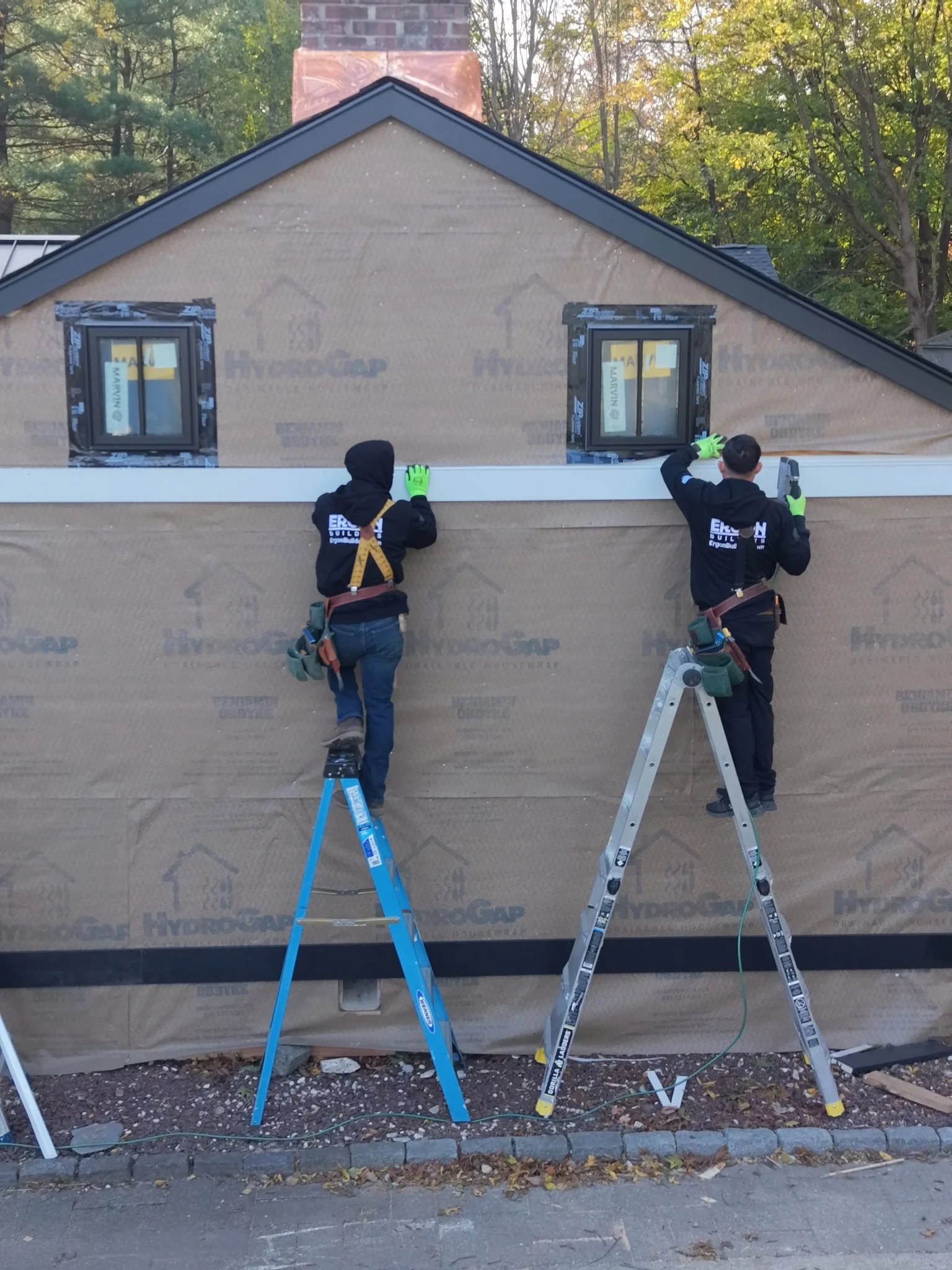 Two construction workers installing a new exterior wall on a house. One is standing on a blue ladder, and the other is on a silver ladder, both working on the wall. The house has two small windows, and the background shows trees with fall foliage.