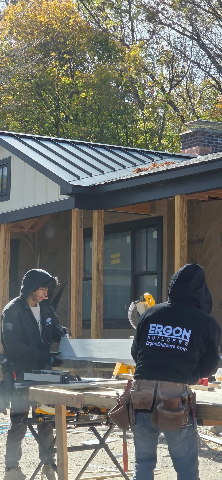Two construction workers wearing black hoodies working on a house construction site, with a metal roof installed, wooden beams, and trees in the background.