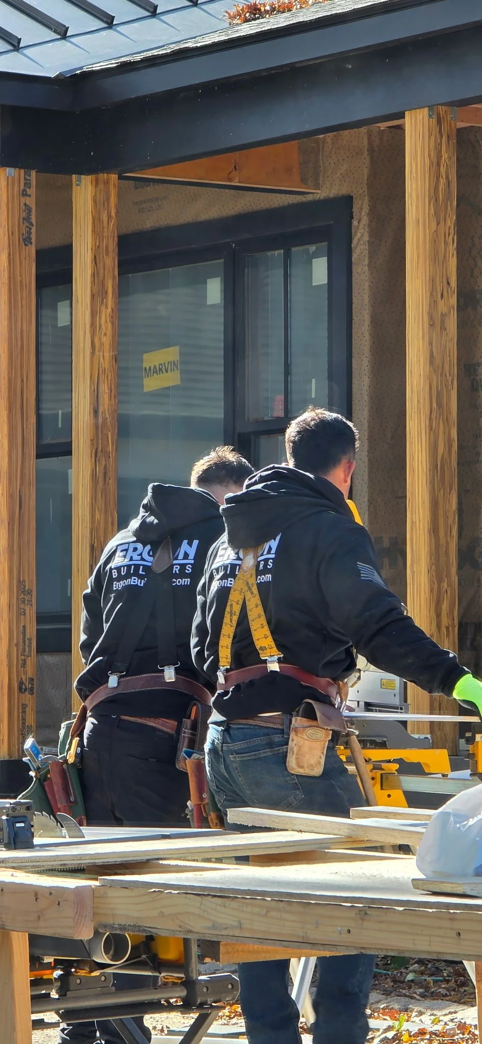 Two construction workers wearing black hoodies with 'Ergon Builders' logos, safety harnesses, and tool belts working on a building site with wooden framing.