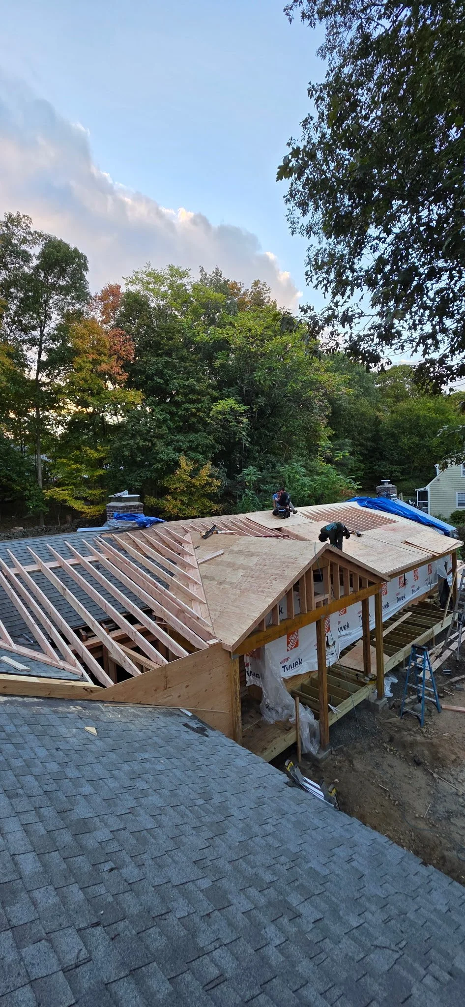Construction workers are building a new roof on a house, with wooden framing and plywood in place, surrounded by trees and other houses.