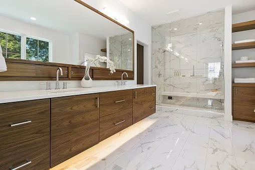 Modern bathroom with wooden vanity, white countertops, large mirror, marble shower, and open shelving with towels.