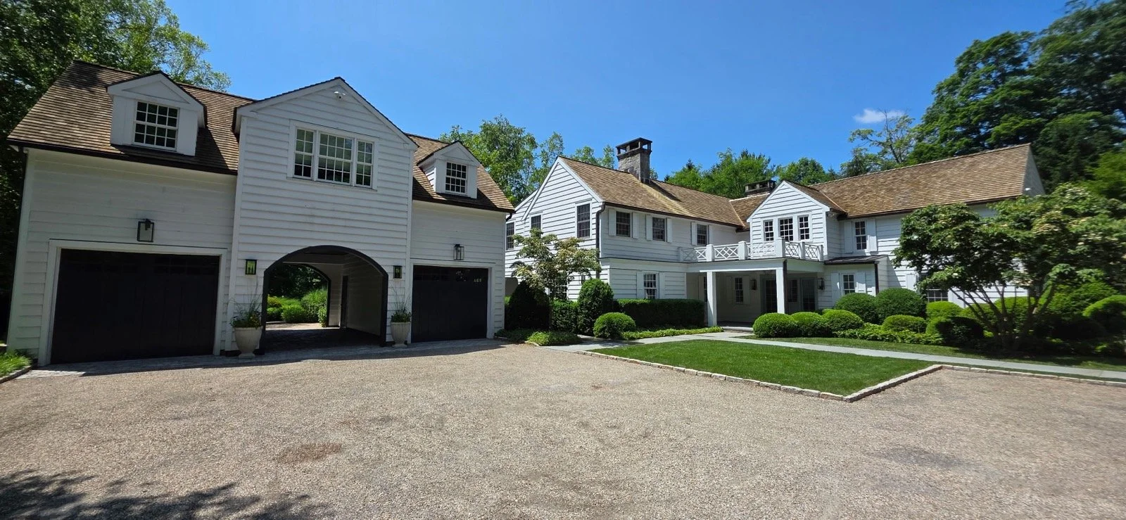 A large white house with a garage, surrounded by greenery and a gravel driveway under a blue sky.