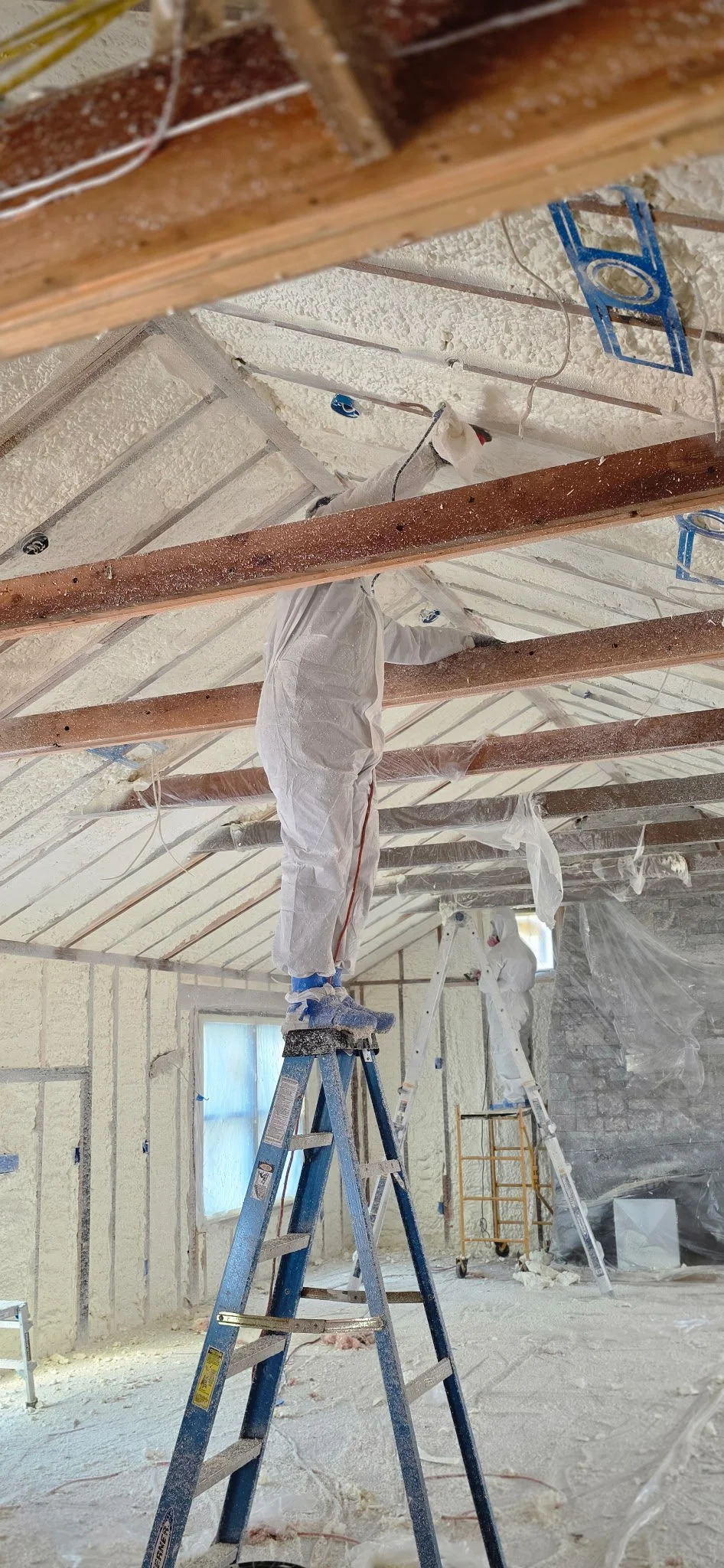 Construction workers in protective suits working on ceiling insulation installation in a room, using ladders and scaffolding.