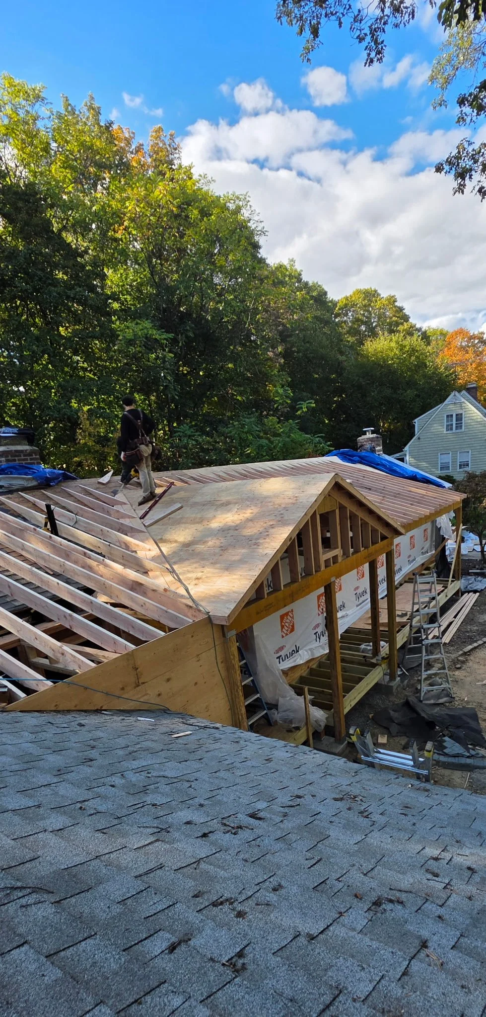 A house under construction with workers installing a new roof, wooden framing, and blue tarps on the roof. Surrounding trees and a neighboring house are visible under a partly cloudy sky.