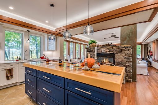 Modern kitchen with blue cabinets, wooden countertop island, pendant lights, large windows with outdoor view, stone fireplace in living area.