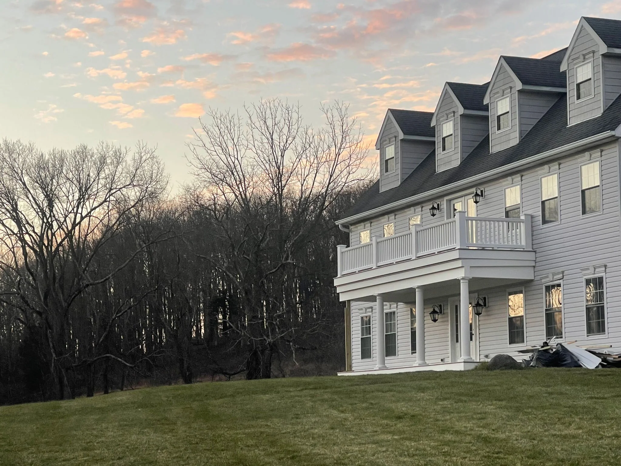 A white multi-story house with a large porch and multiple gabled dormer windows, set against a backdrop of leafless trees and a sunset sky.