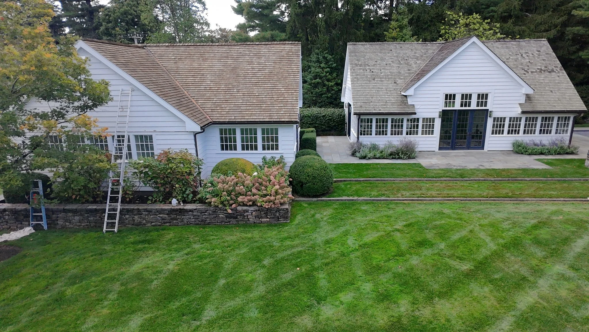 Aerial view of two white houses separated by a stone wall, with manicured lawn, bushes, and trees, in a lush green yard.