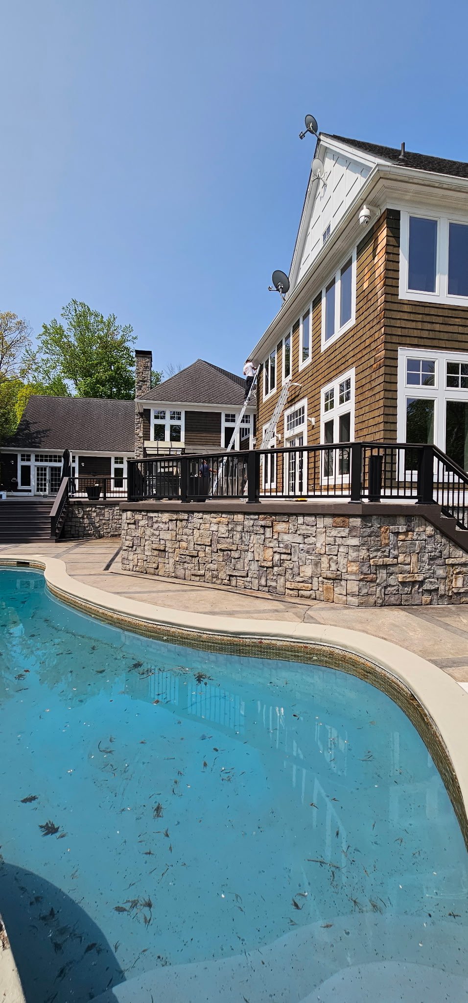 Backyard pool with a stone border and a wooden deck, a house with large windows, and a clear blue sky.