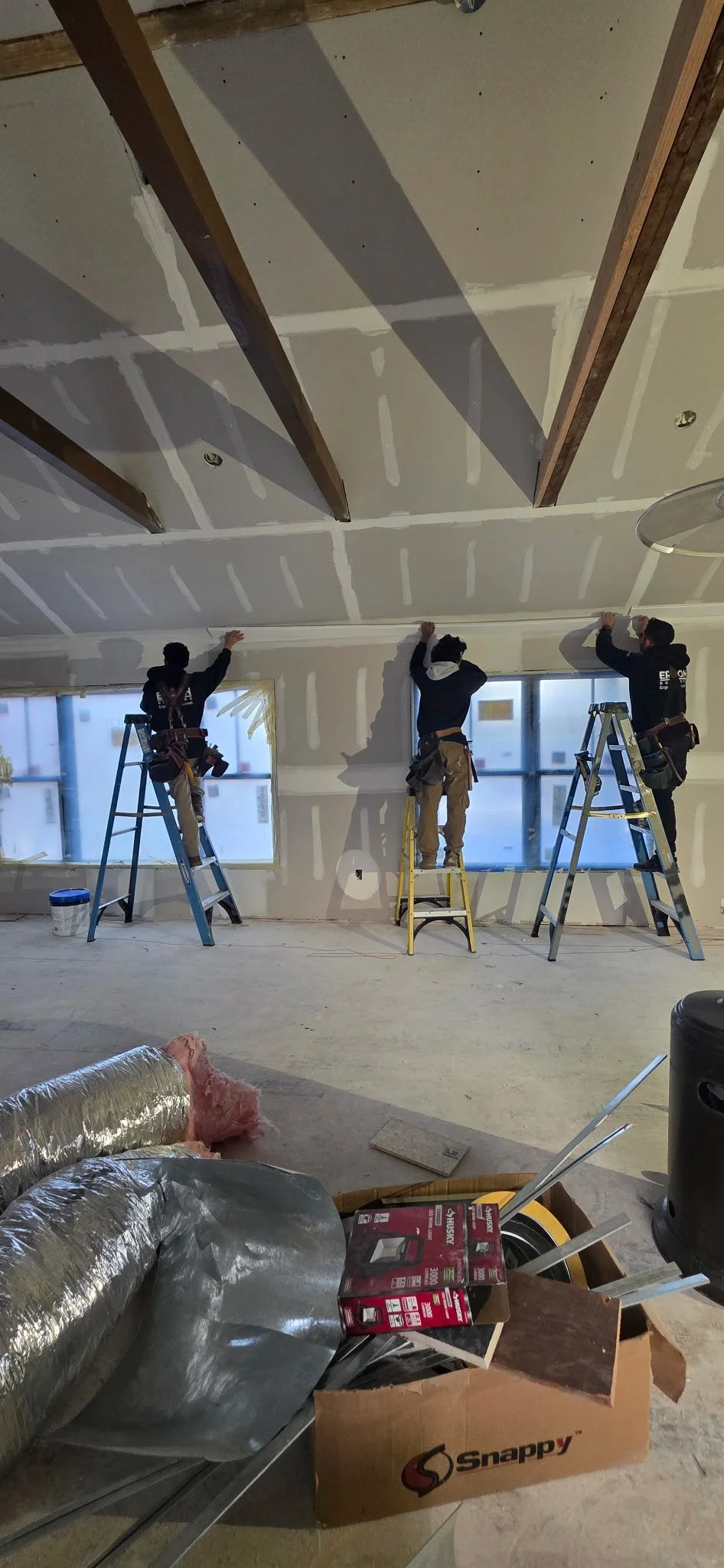 Three workers on ladders installing or finishing drywall on a ceiling. The room has large windows and construction supplies in the foreground.