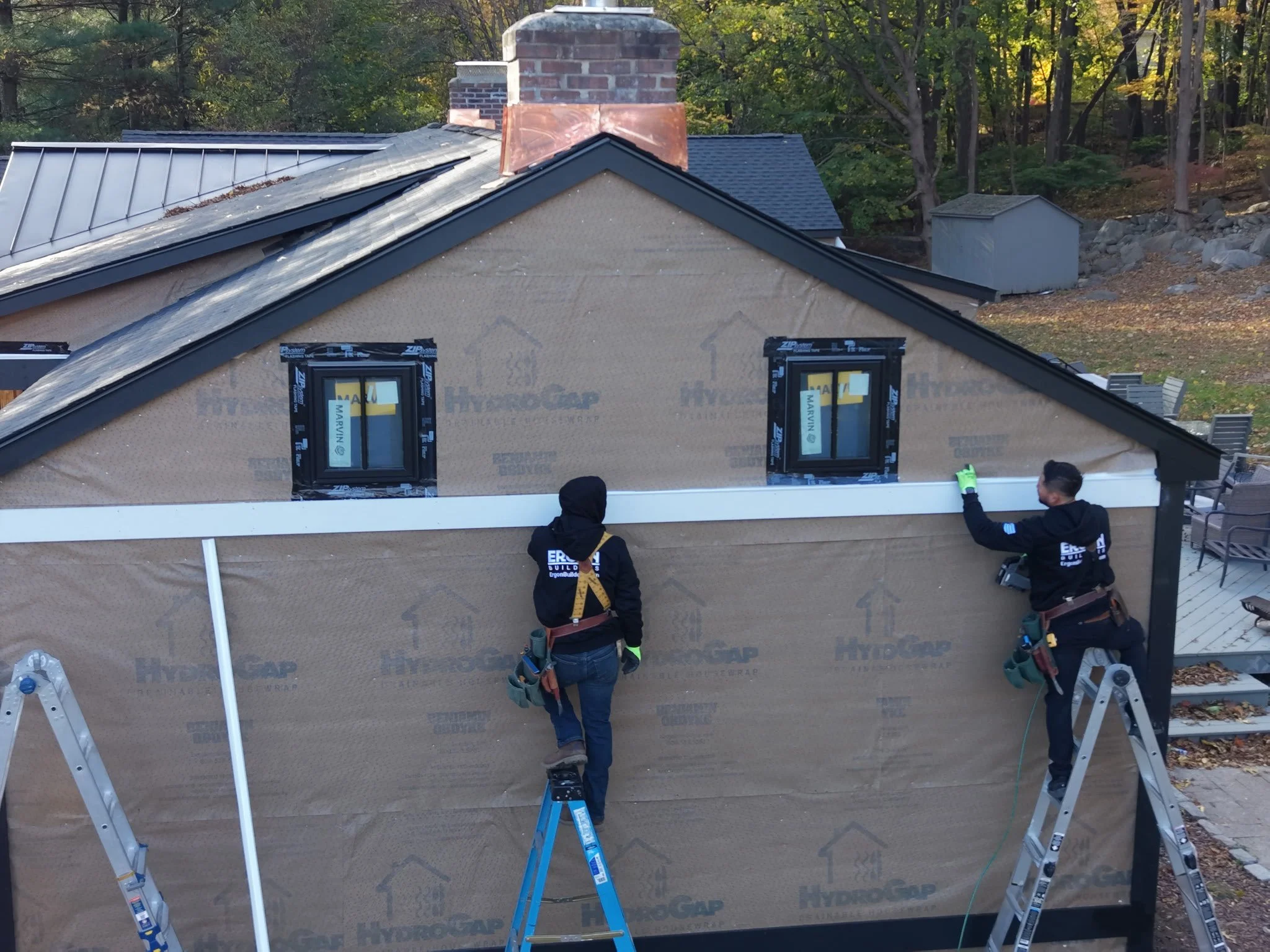 Two construction workers installing exterior siding on a house, standing on ladders, with black hoodies and tool belts, working on the side of the house near two small windows.