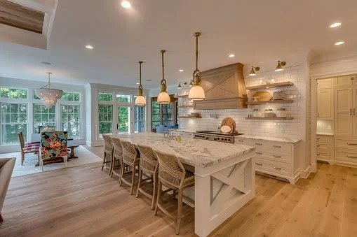 Open-concept kitchen with white cabinetry, marble island, pendant lighting, and a dining area with large windows and a floral armchair.