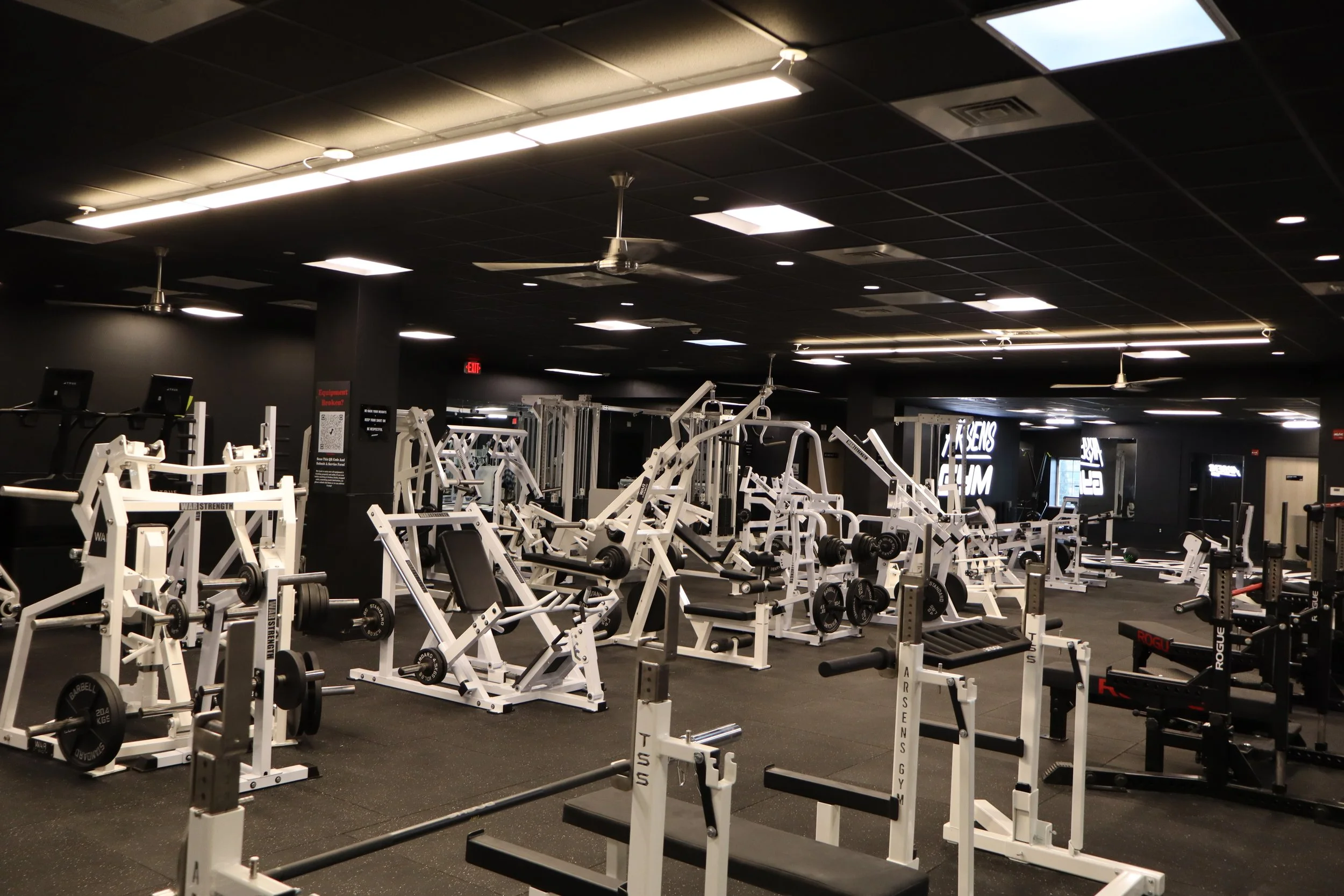 Empty gym with various white weightlifting machines and equipment on a black floor, black walls, and a ceiling with bright white lights.