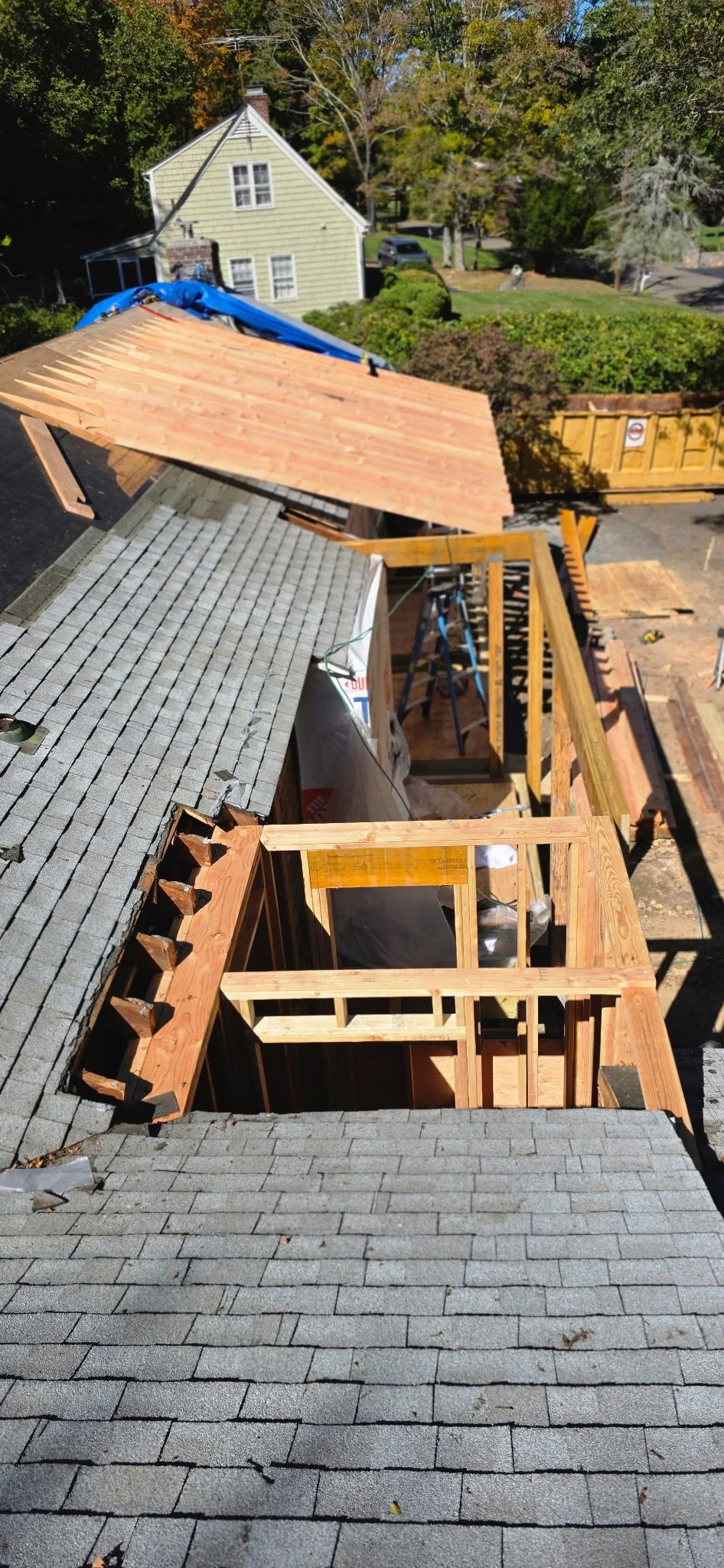 Construction site on a residential roof with wooden framing and partial roof decking, surrounded by trees and neighboring houses.
