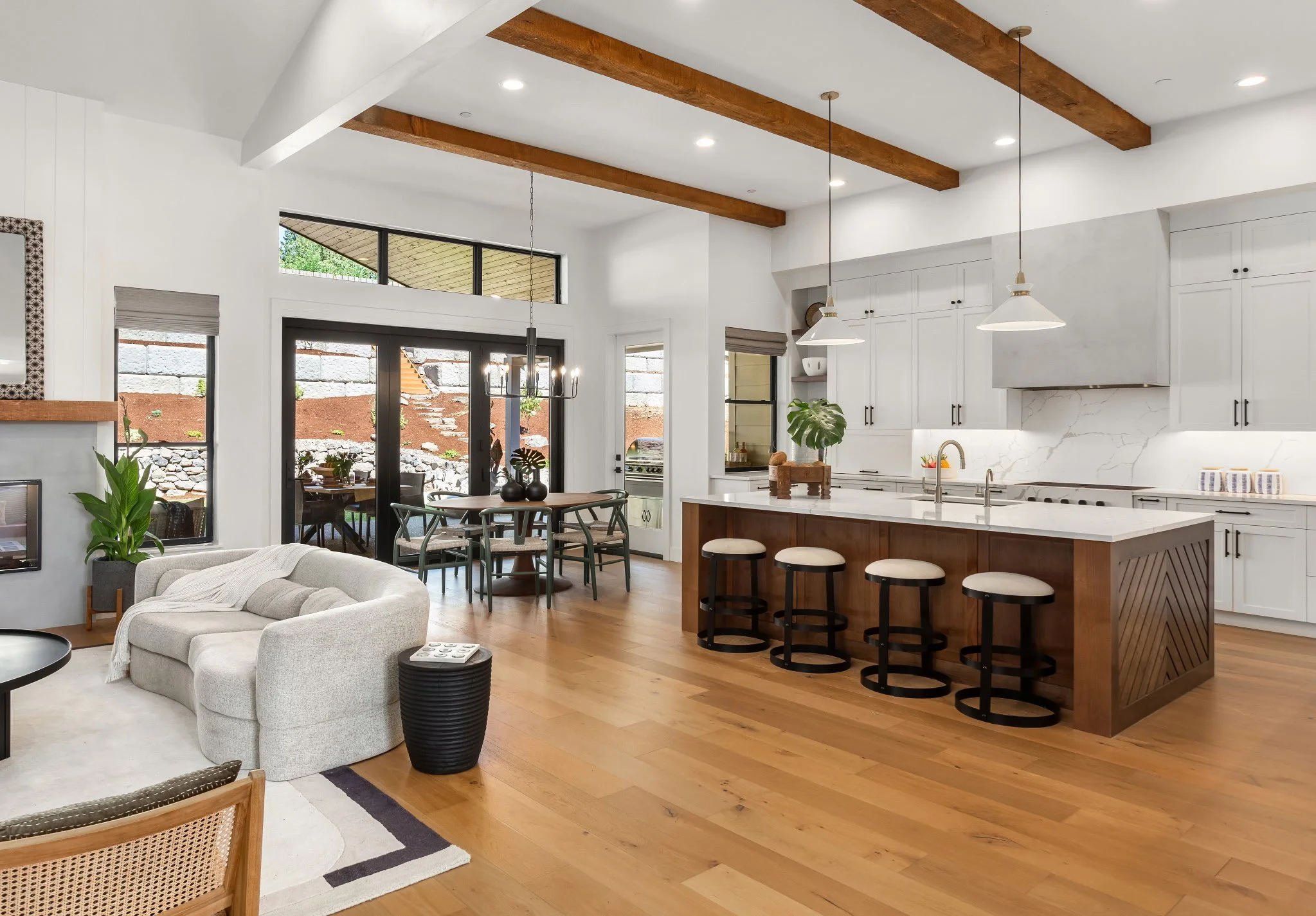 Open-concept kitchen and living room with white cabinetry, marble backsplash, wooden beams, and hardwood floors, featuring a kitchen island with four stools, a dining table with chairs, and a beige sofa.
