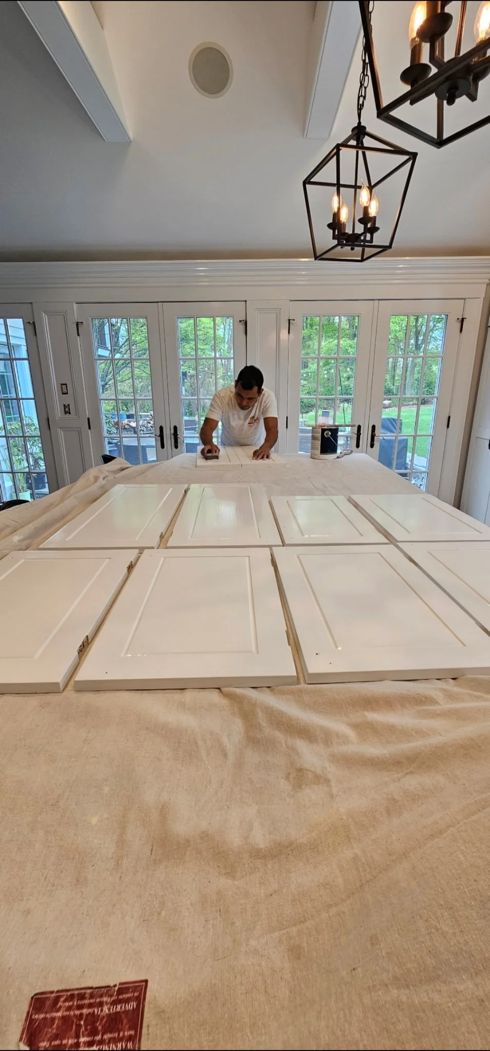 A man working on painting white cabinet doors at a large kitchen table in a room with large glass doors and windows, hanging black metal chandeliers, and green trees outside.