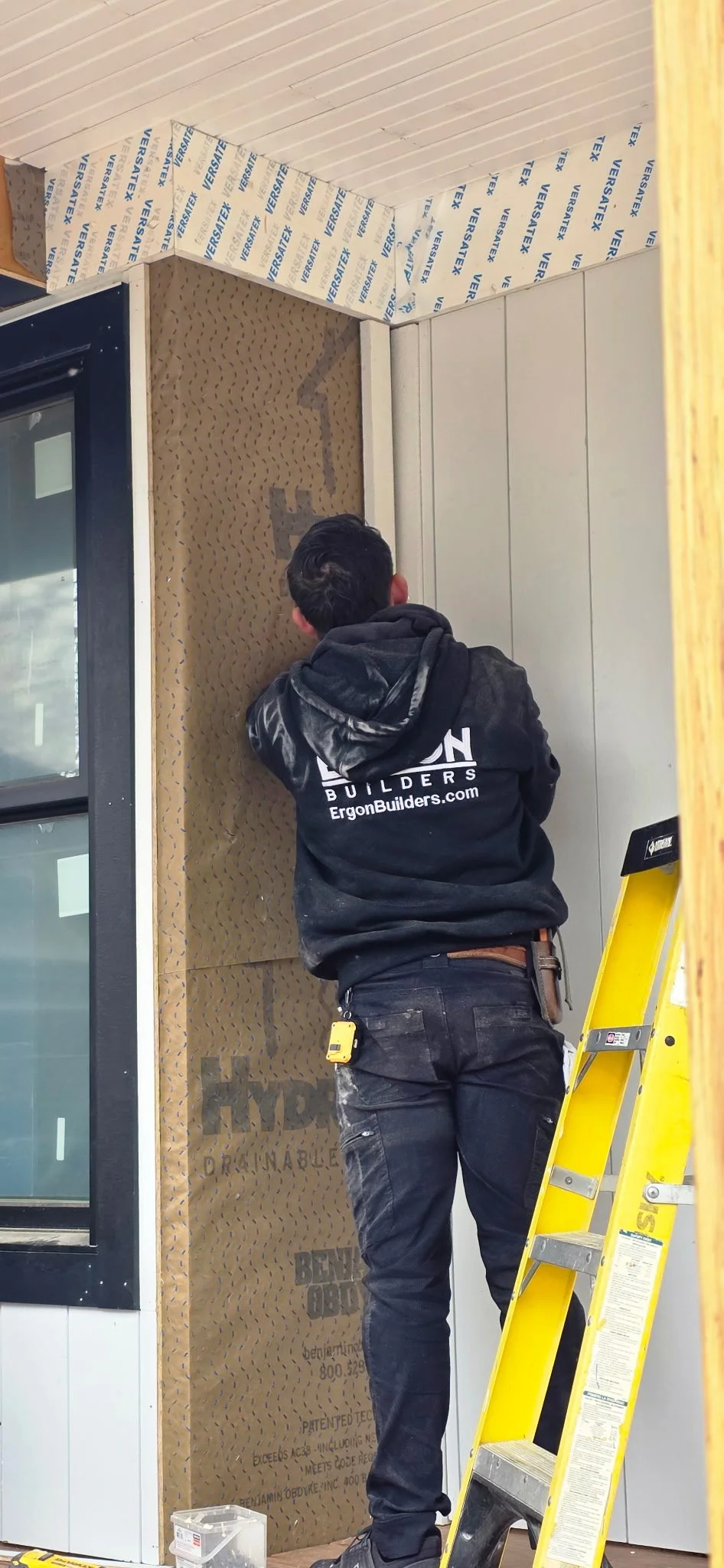 A worker installing insulation behind a wall in a building, with a yellow ladder nearby.