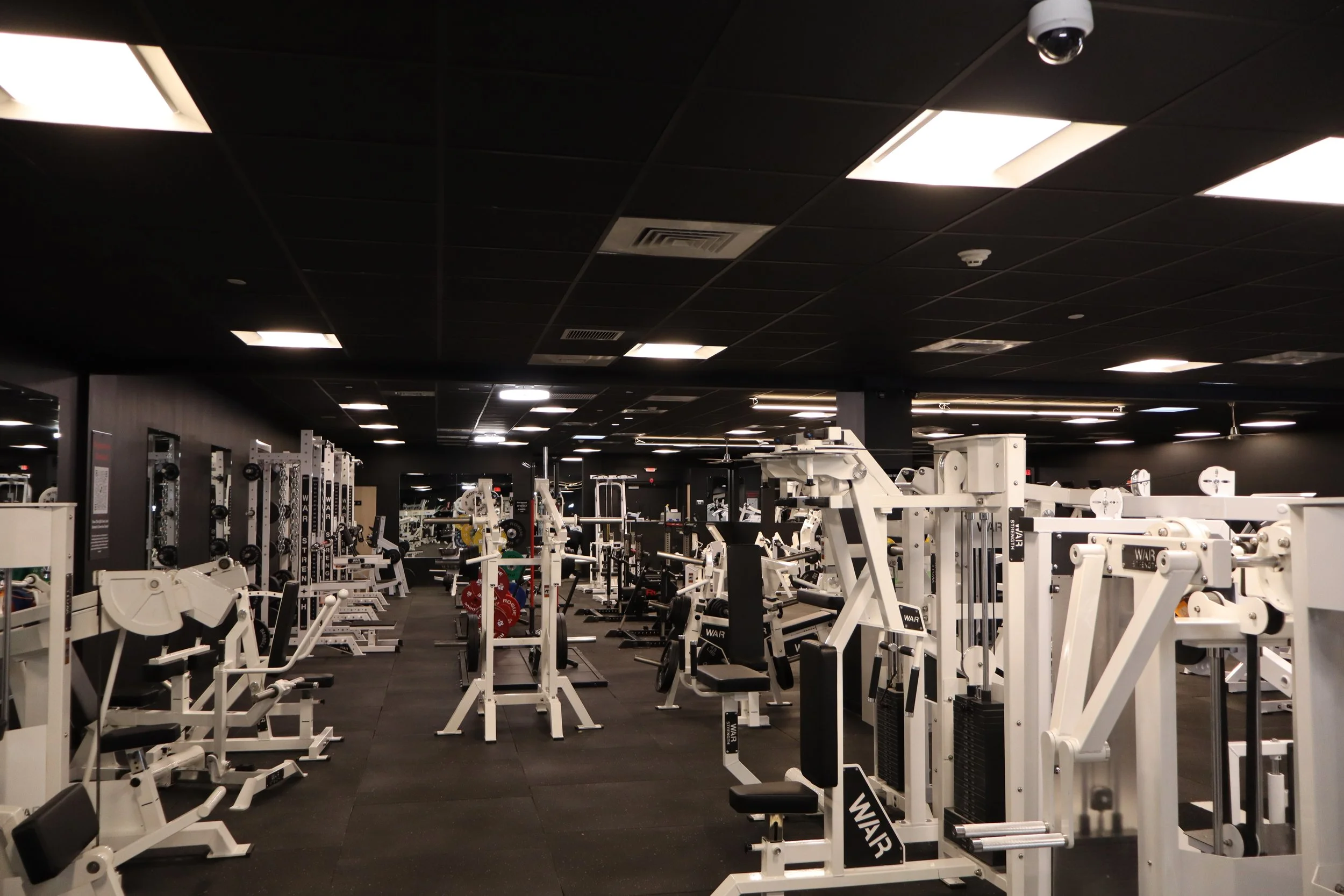 Empty gym with various weight machines and equipment, black walls, black ceiling, and bright overhead lights.