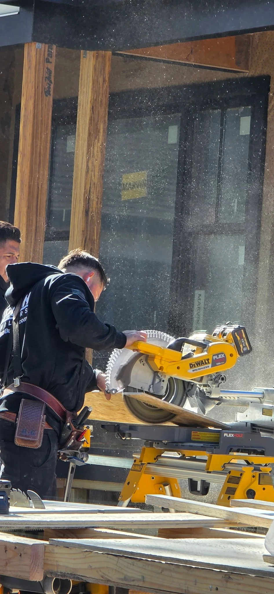 Two workers operating a yellow DeWalt cordless circular saw cutting wood on a construction site, with dust and sunlight. The background shows a black and brown building exterior with attached windows.